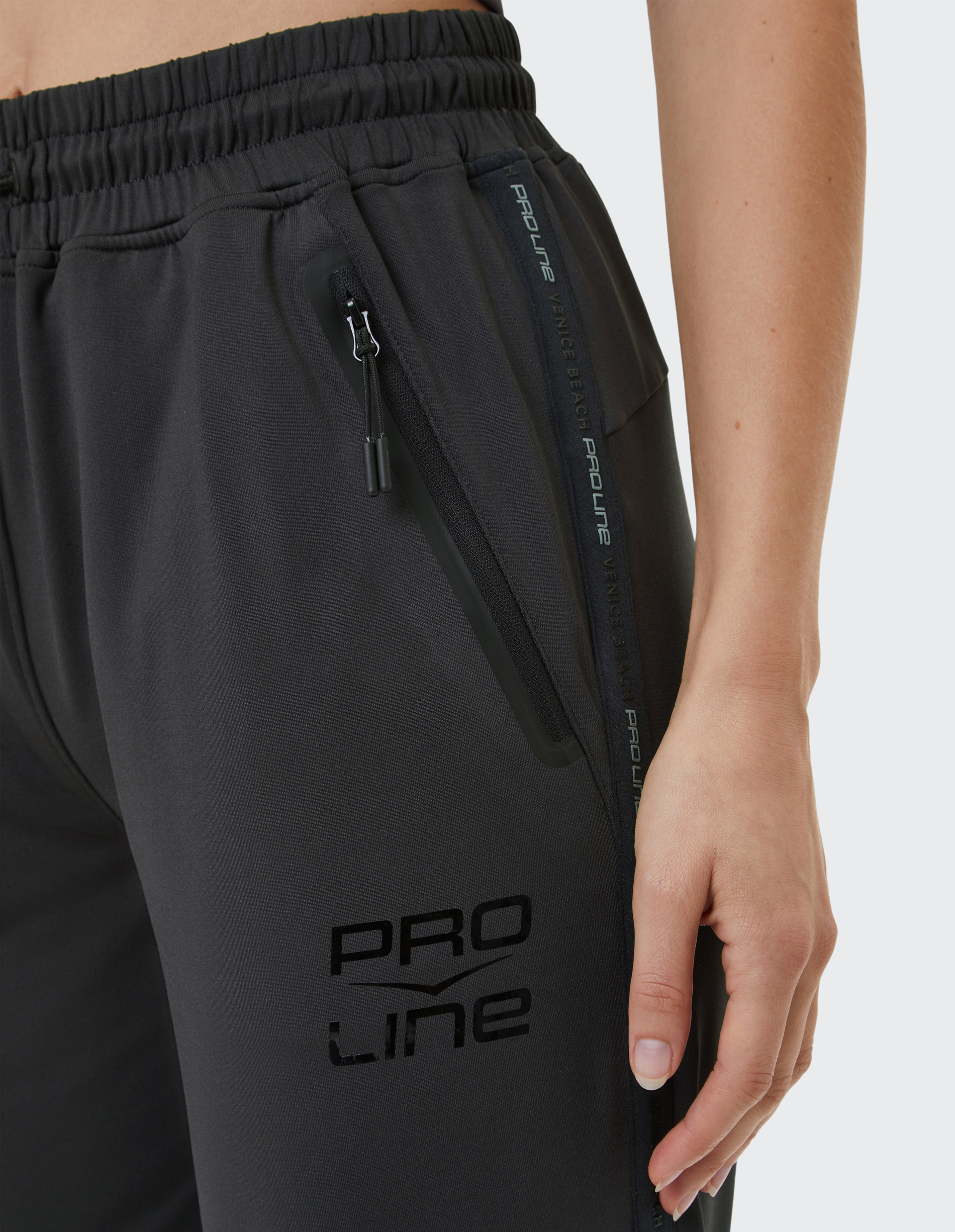 Schwarze Jogginghose mit elastischem Bund, seitlicher Reißverschlusstasche und "PRO LINE"-Aufdruck, Hand neben der Tasche.
