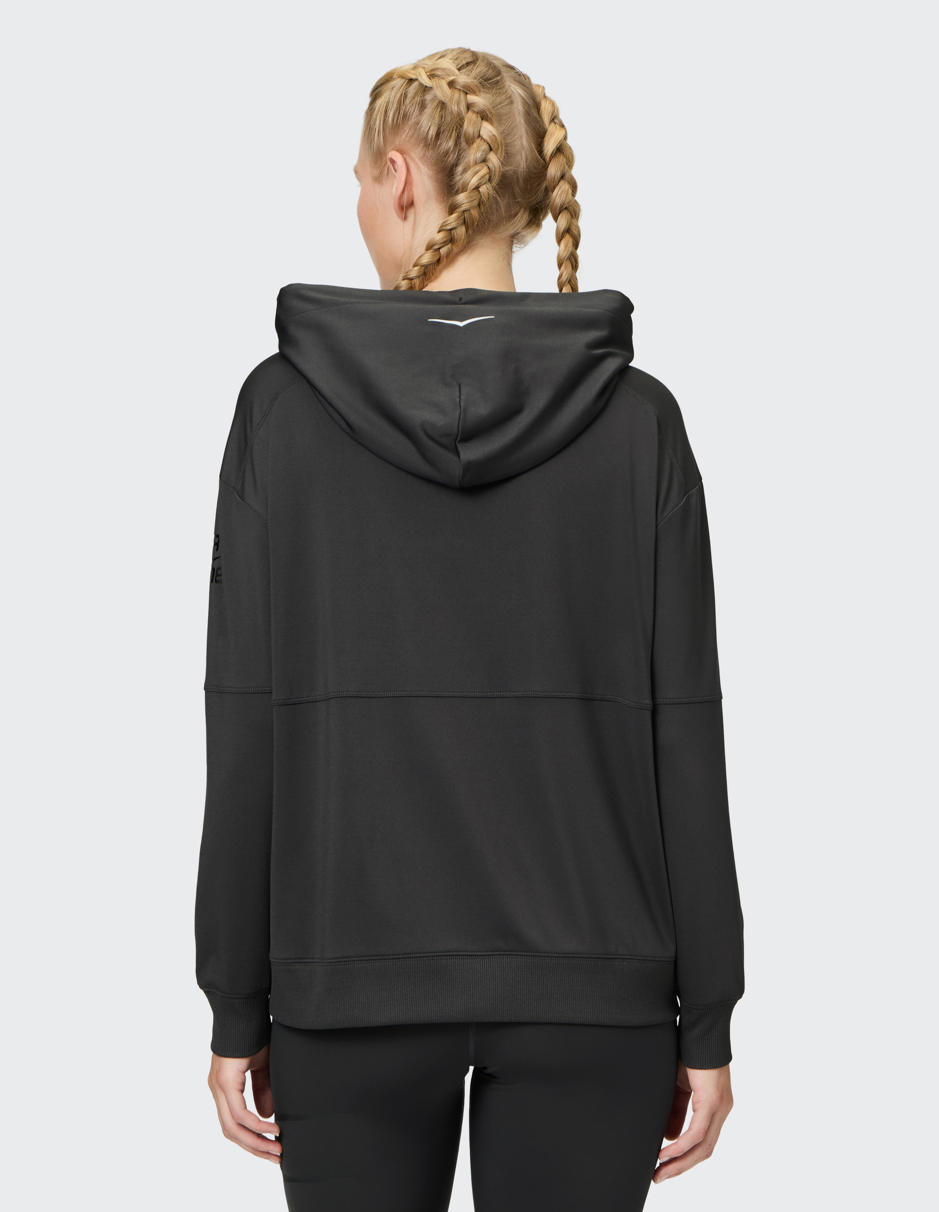 Person mit blonden Zöpfen trägt einen schwarzen Hoodie mit Kapuze und steht mit dem Rücken zur Kamera vor grauem Hintergrund.