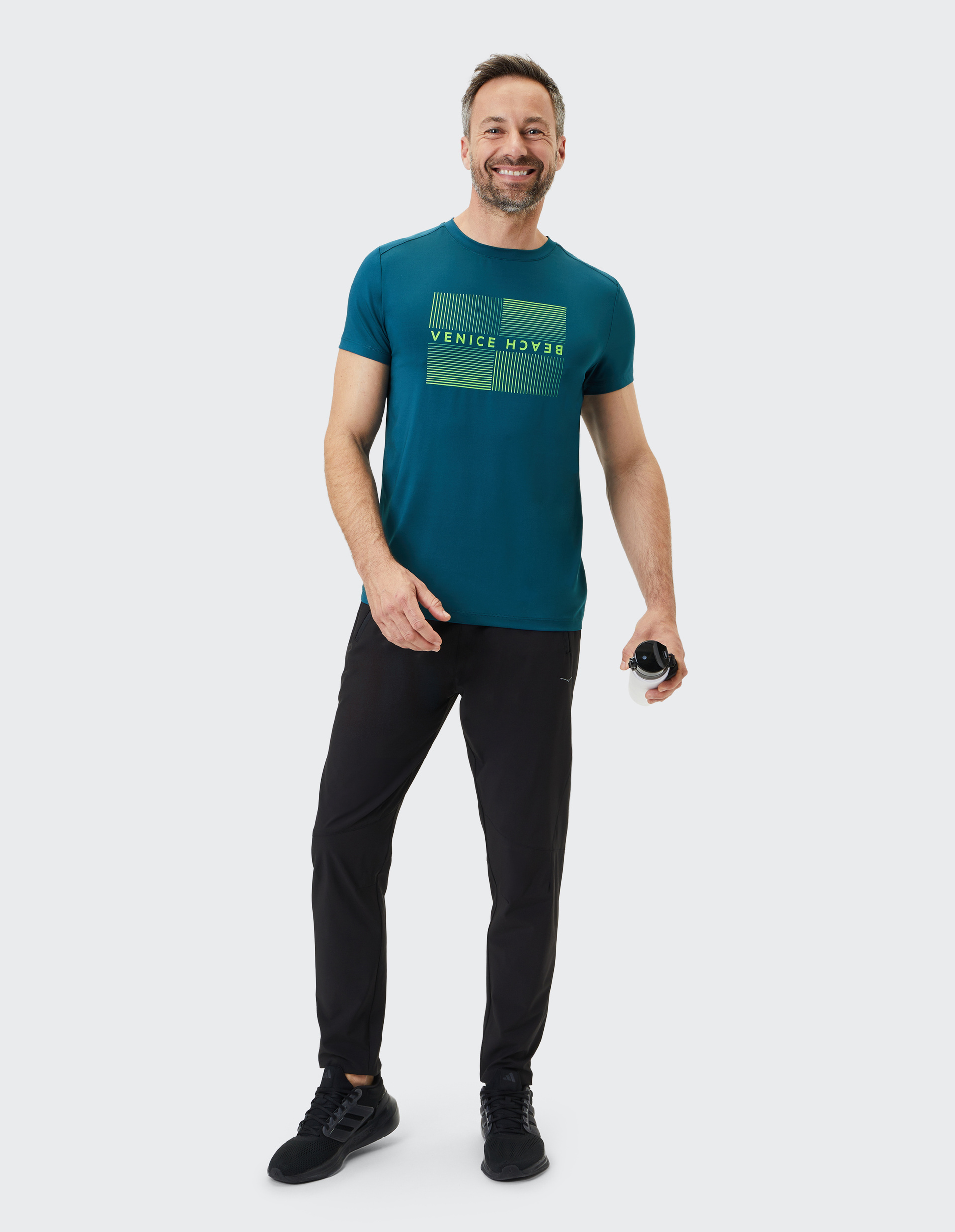 Mann in blauem T-Shirt mit "Venice Beach"-Schrift, lächelnd, in schwarzer Hose und Sneakern, hält eine Wasserflasche.