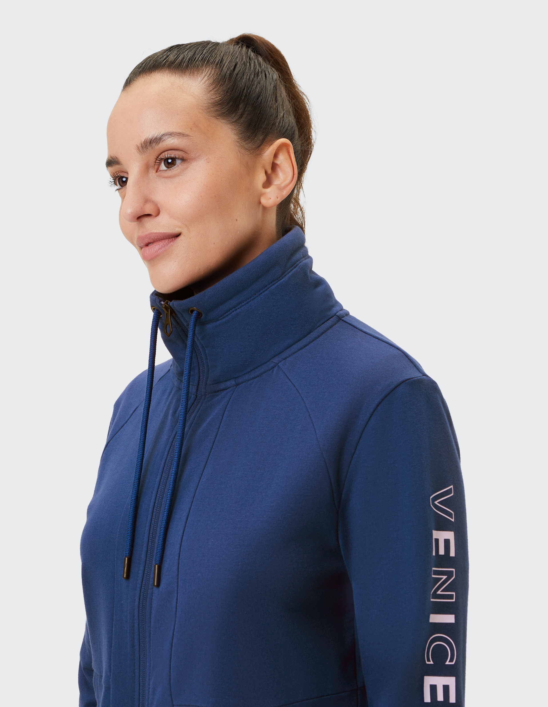 Frau mit Pferdeschwanz trägt eine marineblaue Jacke mit hohem Kragen und "Venice" Schriftzug am Ärmel, lächelnd zur Seite blickend.