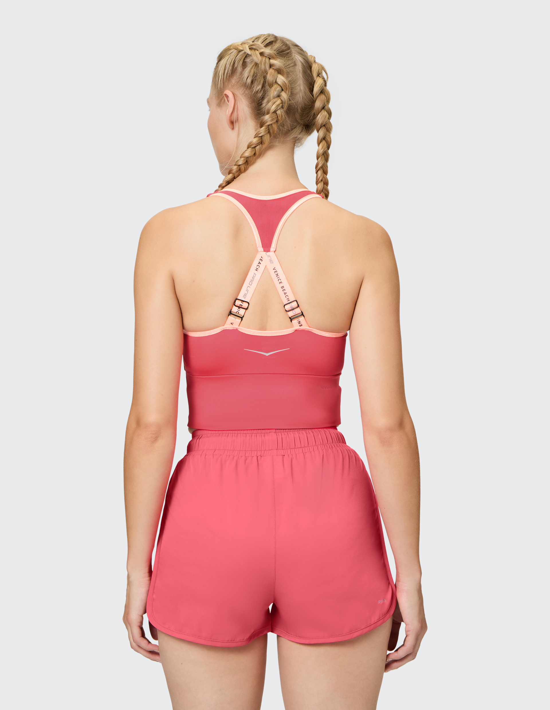 Frau mit blonden Zöpfen trägt ein pinkes Sporttop und passende Shorts, Rückansicht, auf neutralem Hintergrund.