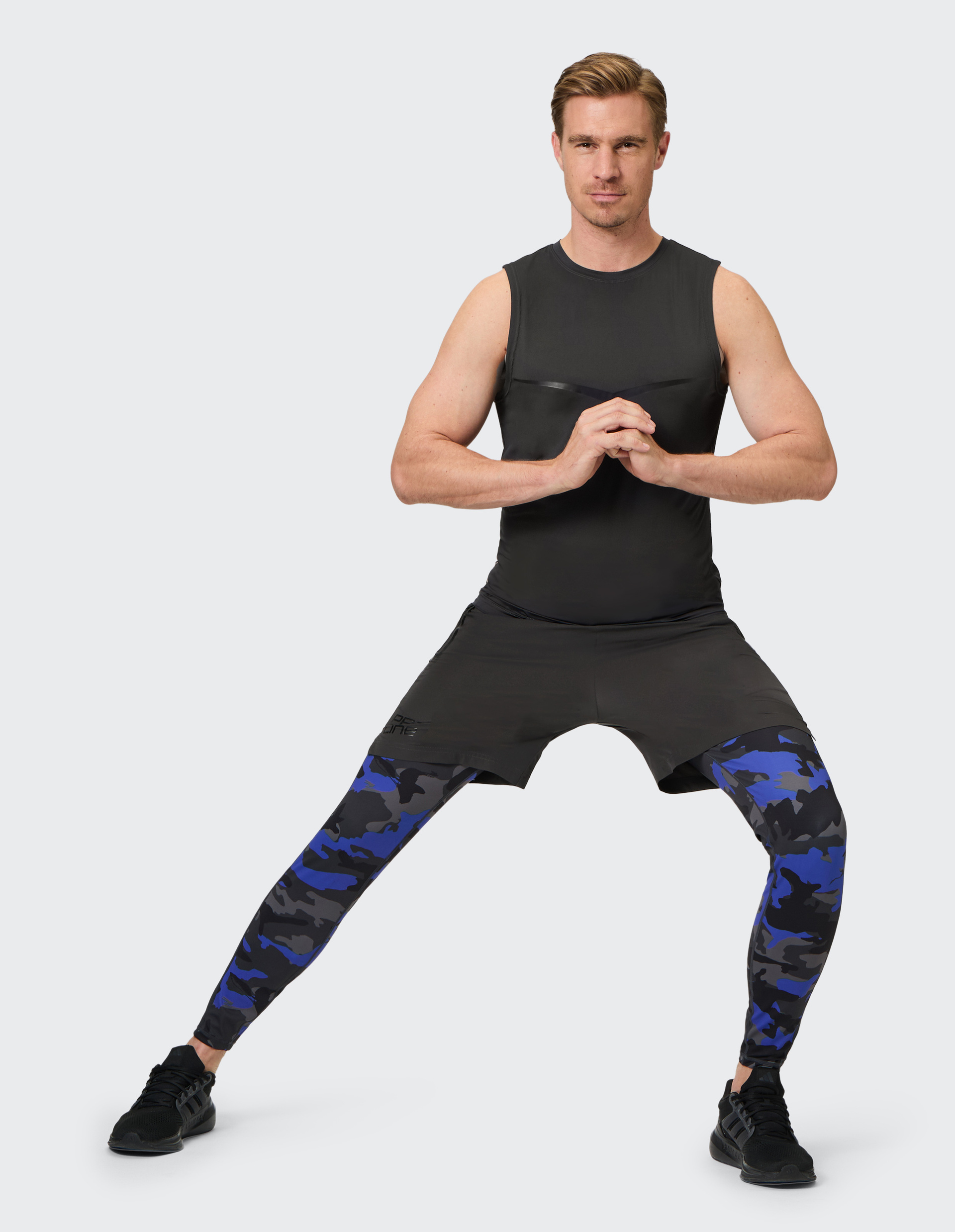 Ein Mann in schwarzem Sportoutfit mit Camouflage-Leggings steht in breiter Ausfallschrittposition vor weißem Hintergrund.