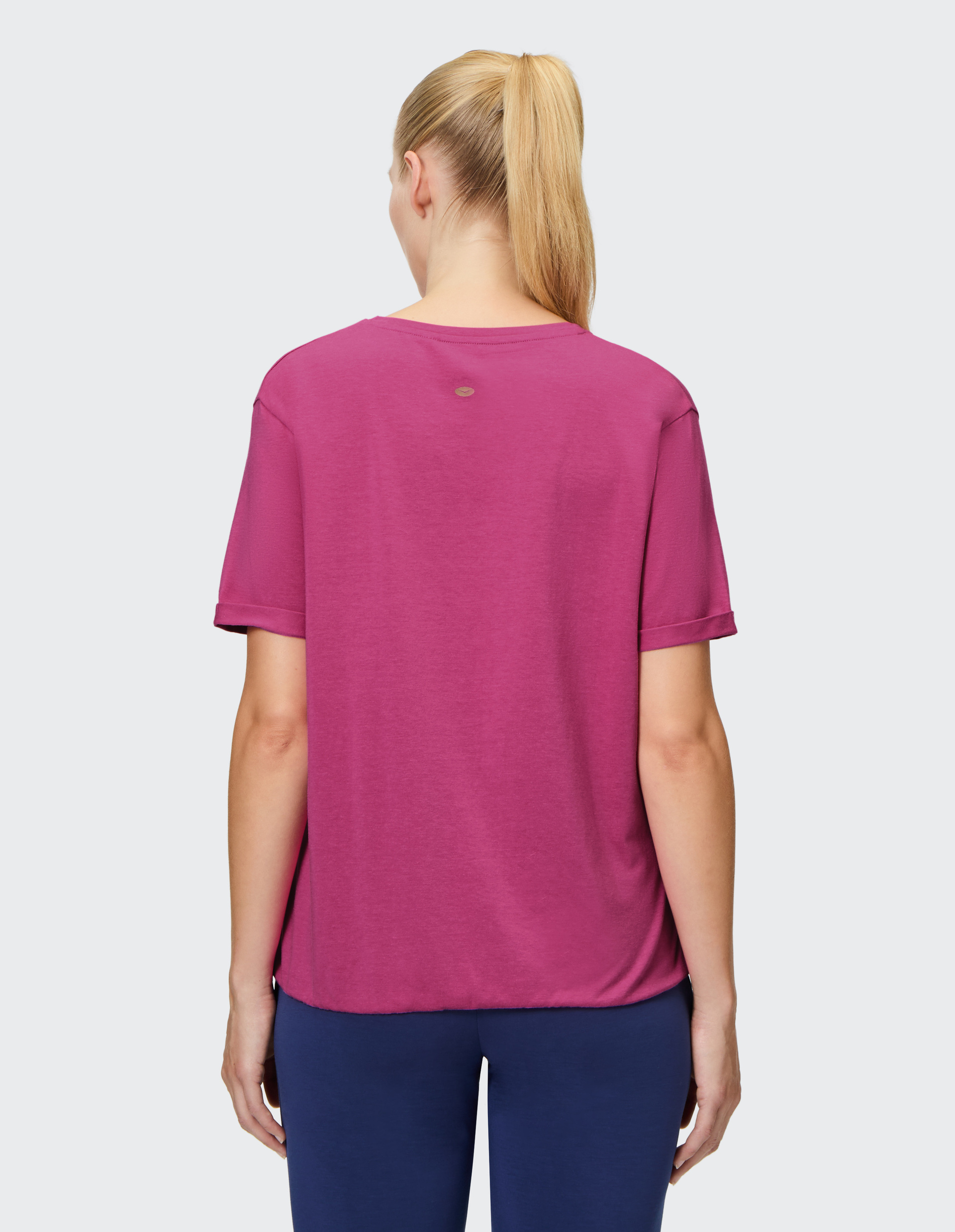 Frau von hinten in fuchsiafarbenem T-Shirt und blauer Hose, blondes Haar zu einem Pferdeschwanz gebunden, auf hellem Hintergrund.