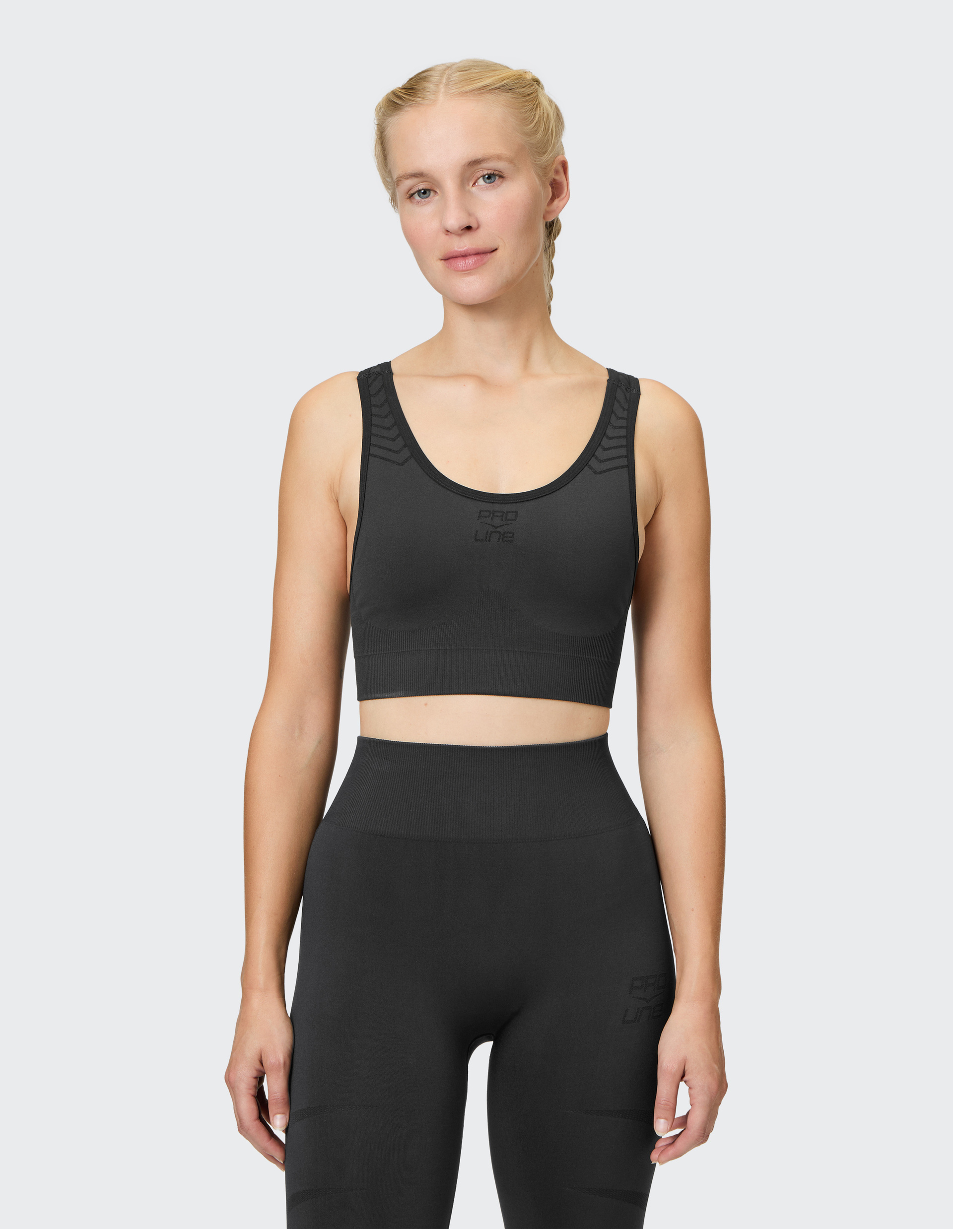Eine Person mit blonden Haaren trägt ein schwarzes, ärmelloses Sport-Oberteil und passende Leggings vor weißem Hintergrund.