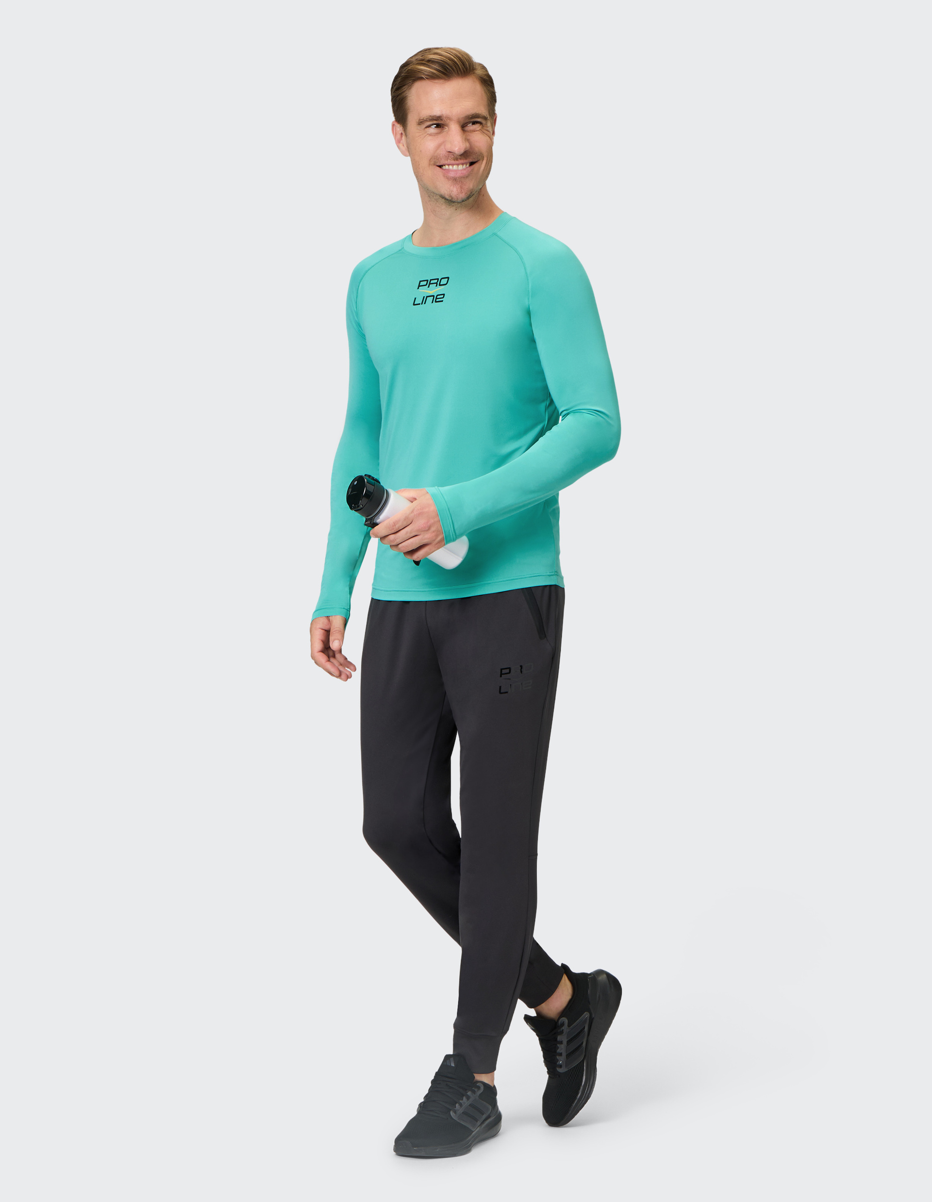 Mann in türkisem Sportshirt und schwarzen Jogginghosen hält eine Wasserflasche und geht lächelnd, grauer Hintergrund.