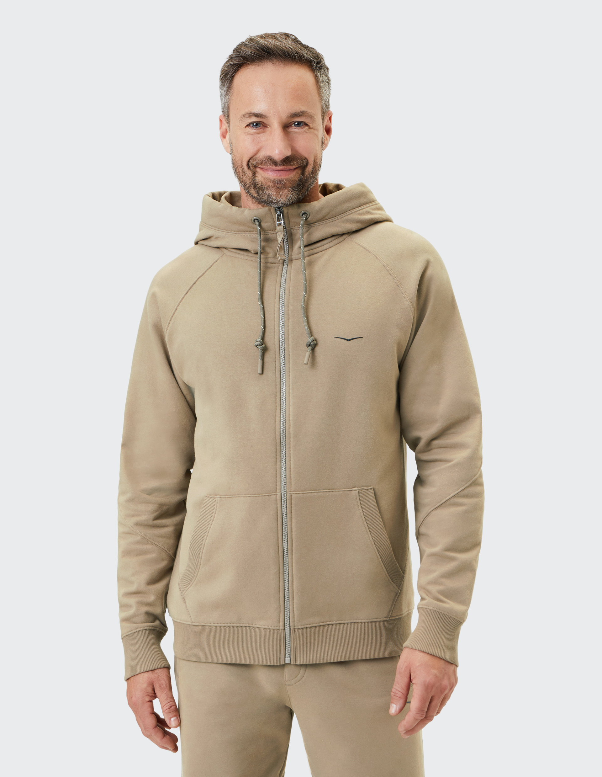 Mann in beiger Kapuzenjacke mit Kordeln und Reißverschluss vor hellem Hintergrund; lächelt leicht und hat braune Haare.
