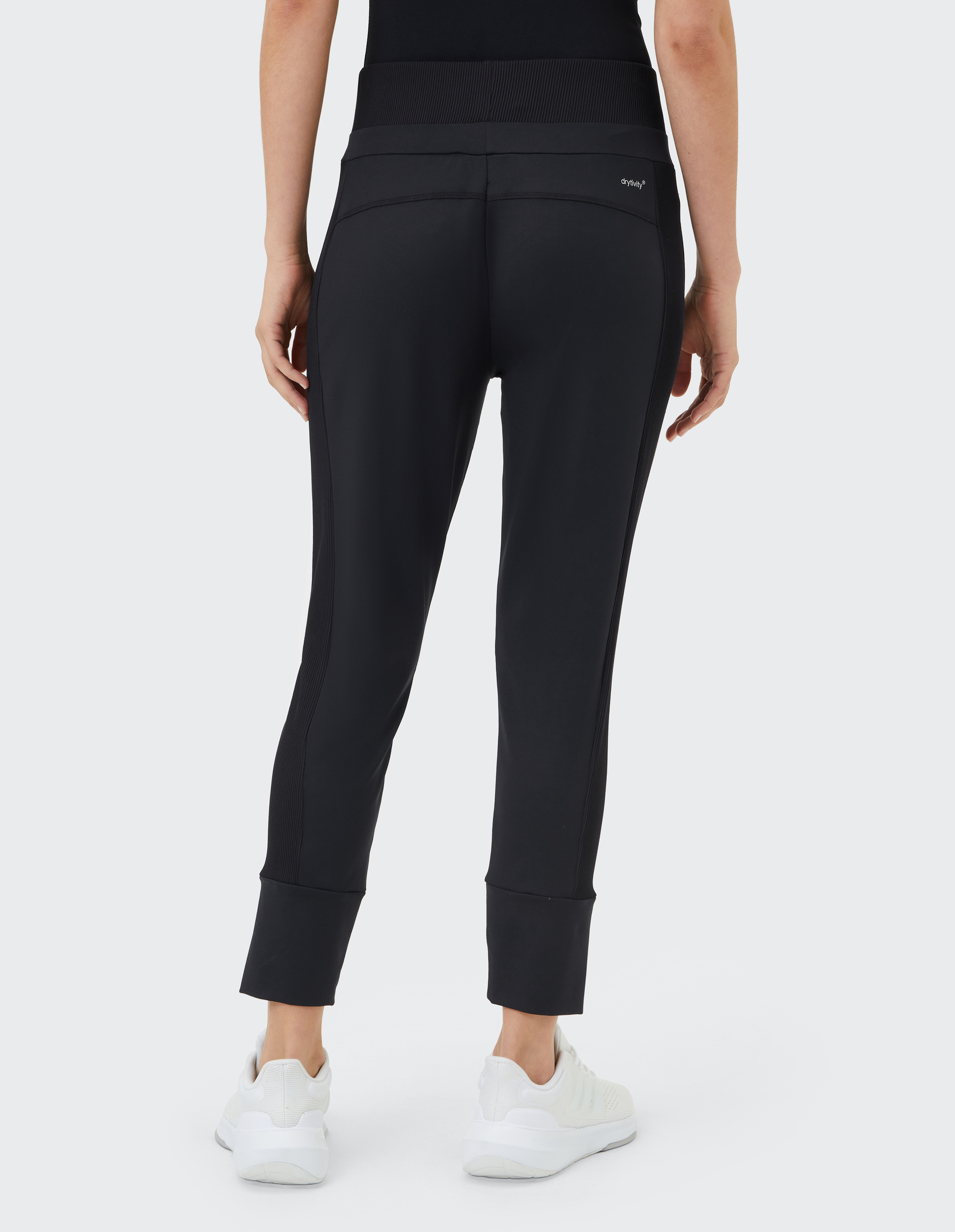 Frau in schwarzer Sporthose mit hohem Bund und weißen Sneakers, von hinten gesehen, vor weißem Hintergrund.