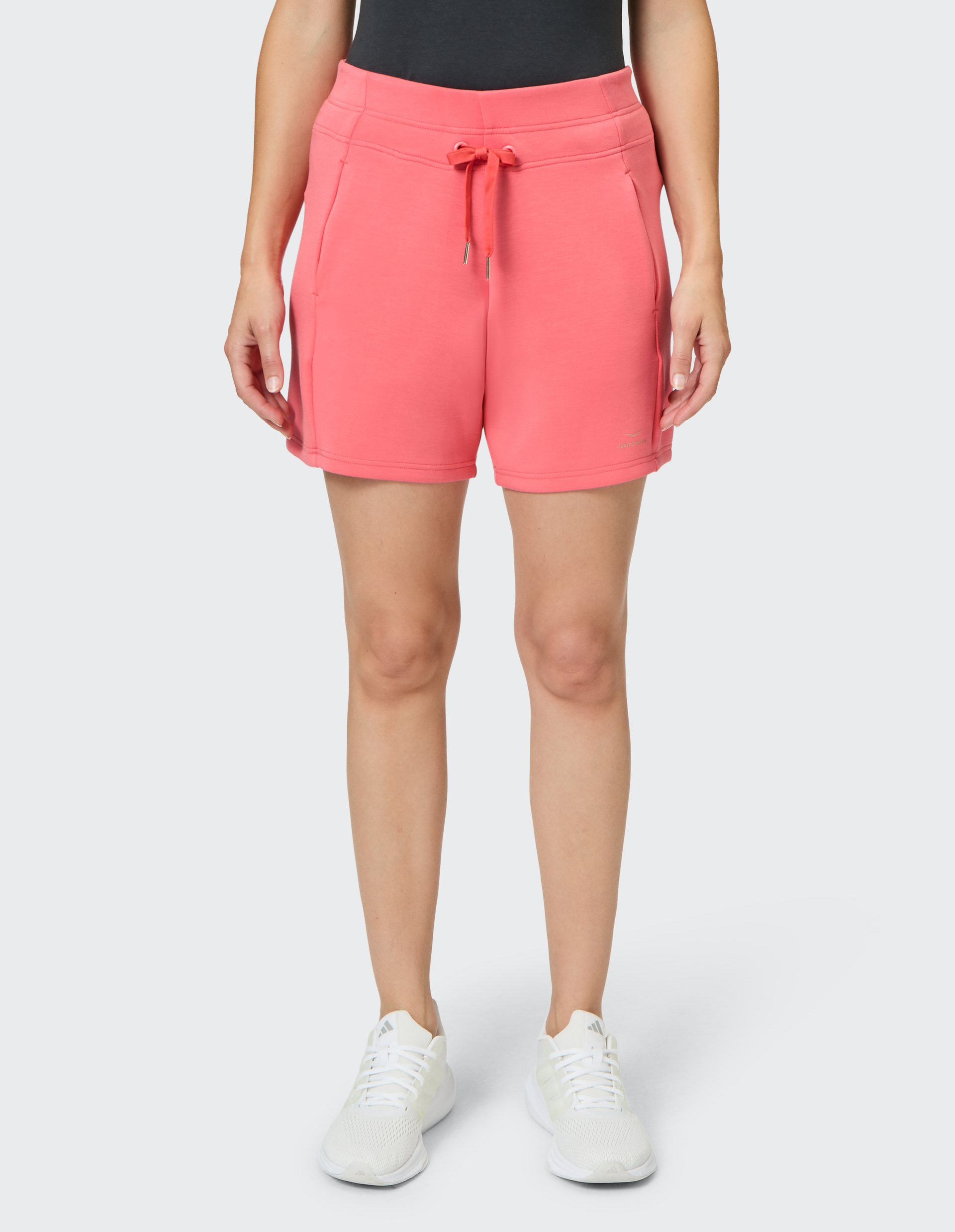 Frau in rosa Shorts und schwarzen Top, steht aufrecht. Weiße Sportschuhe, grauer Hintergrund; sportlicher, lässiger Look.