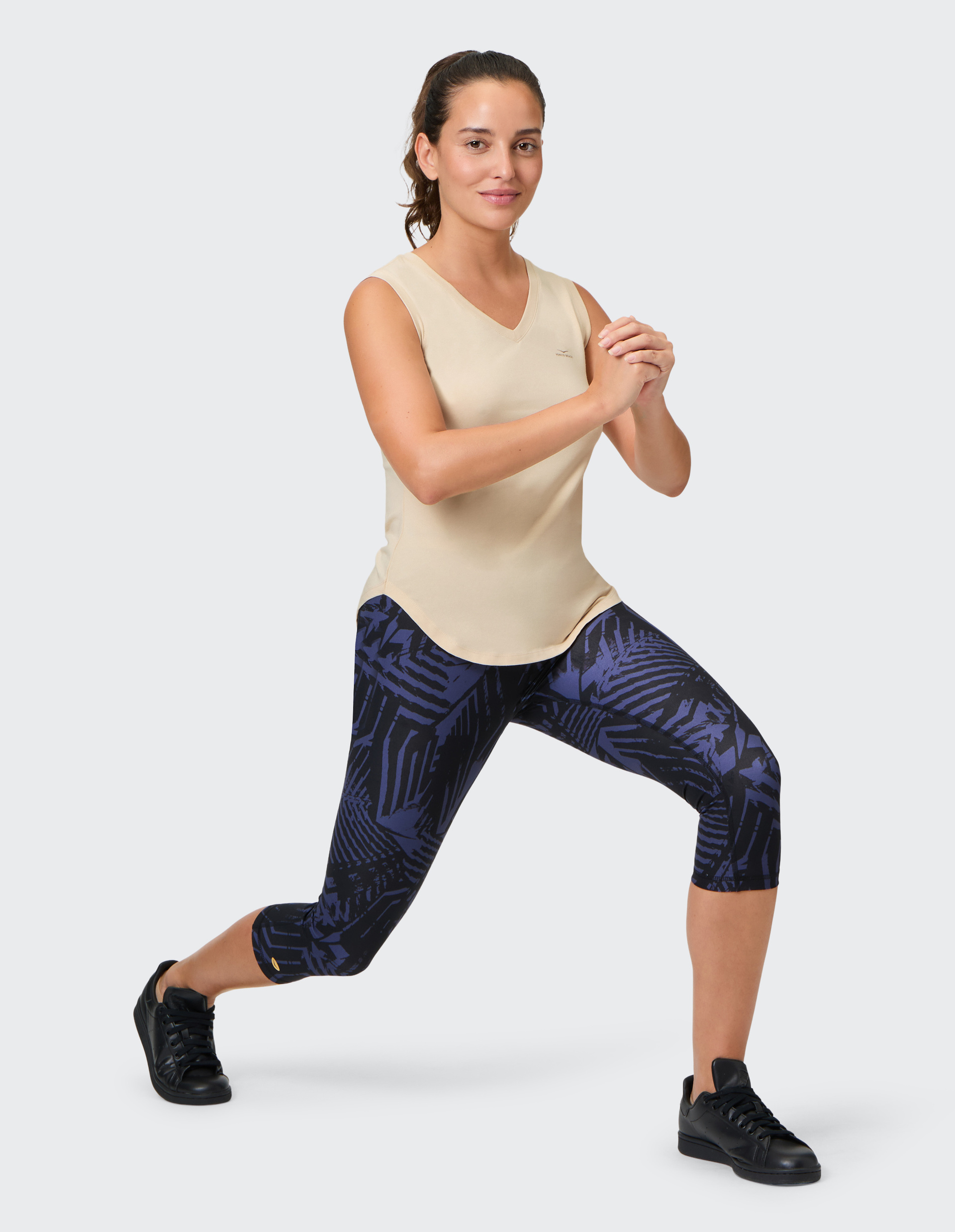 Frau in beige Tanktop und blau-gemusterten Leggings macht seitlichen Ausfallschritt; Hände vor der Brust gefaltet, trägt schwarze Sneaker.