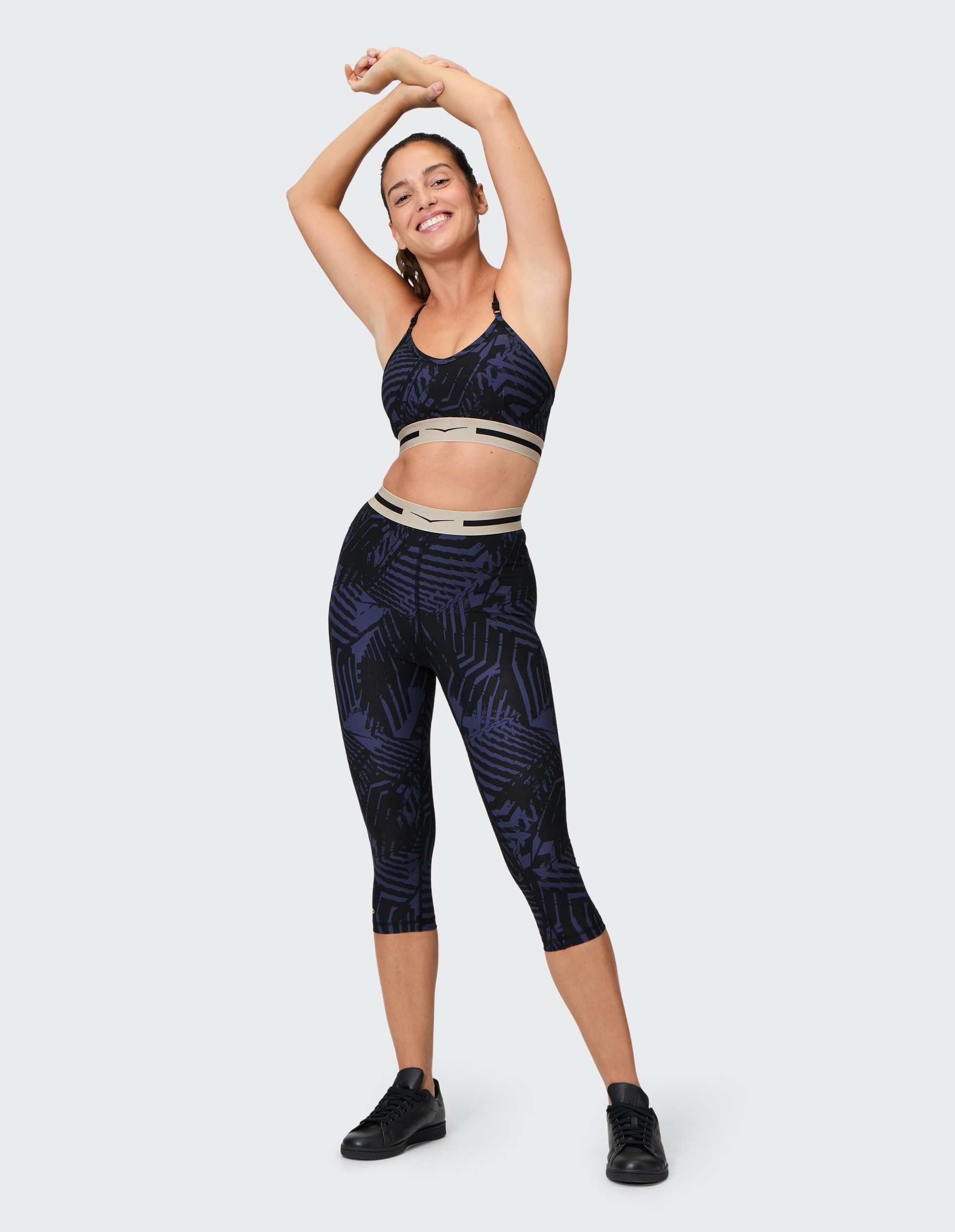 Frau in blau-schwarzem Sportoutfit macht lächelnd Dehnübung mit erhobenen Armen, trägt schwarze Schuhe, steht auf weißem Hintergrund.