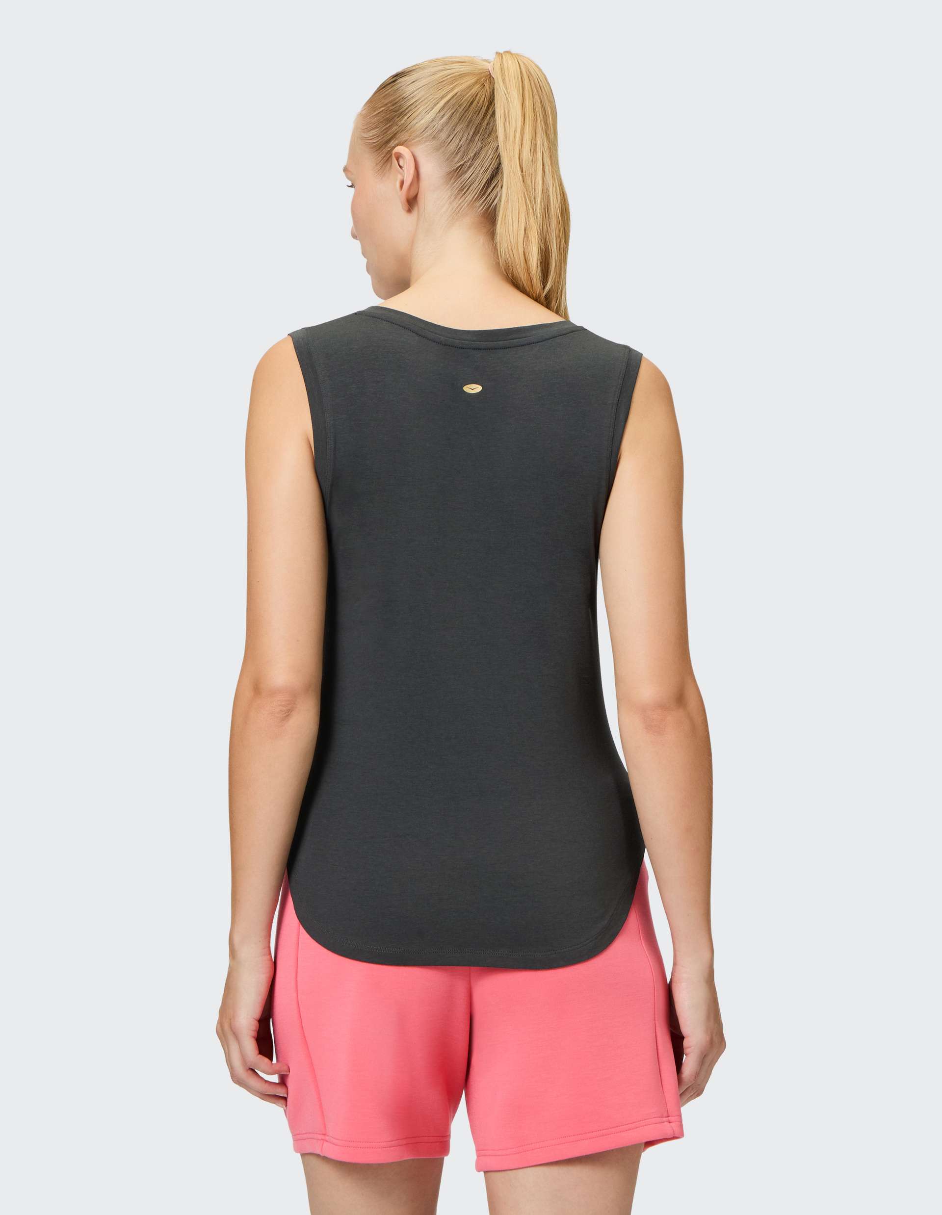 Frau mit blondem Pferdeschwanz trägt ärmelloses, dunkelgraues Oberteil und pinke Shorts, Rückenansicht vor hellem Hintergrund.