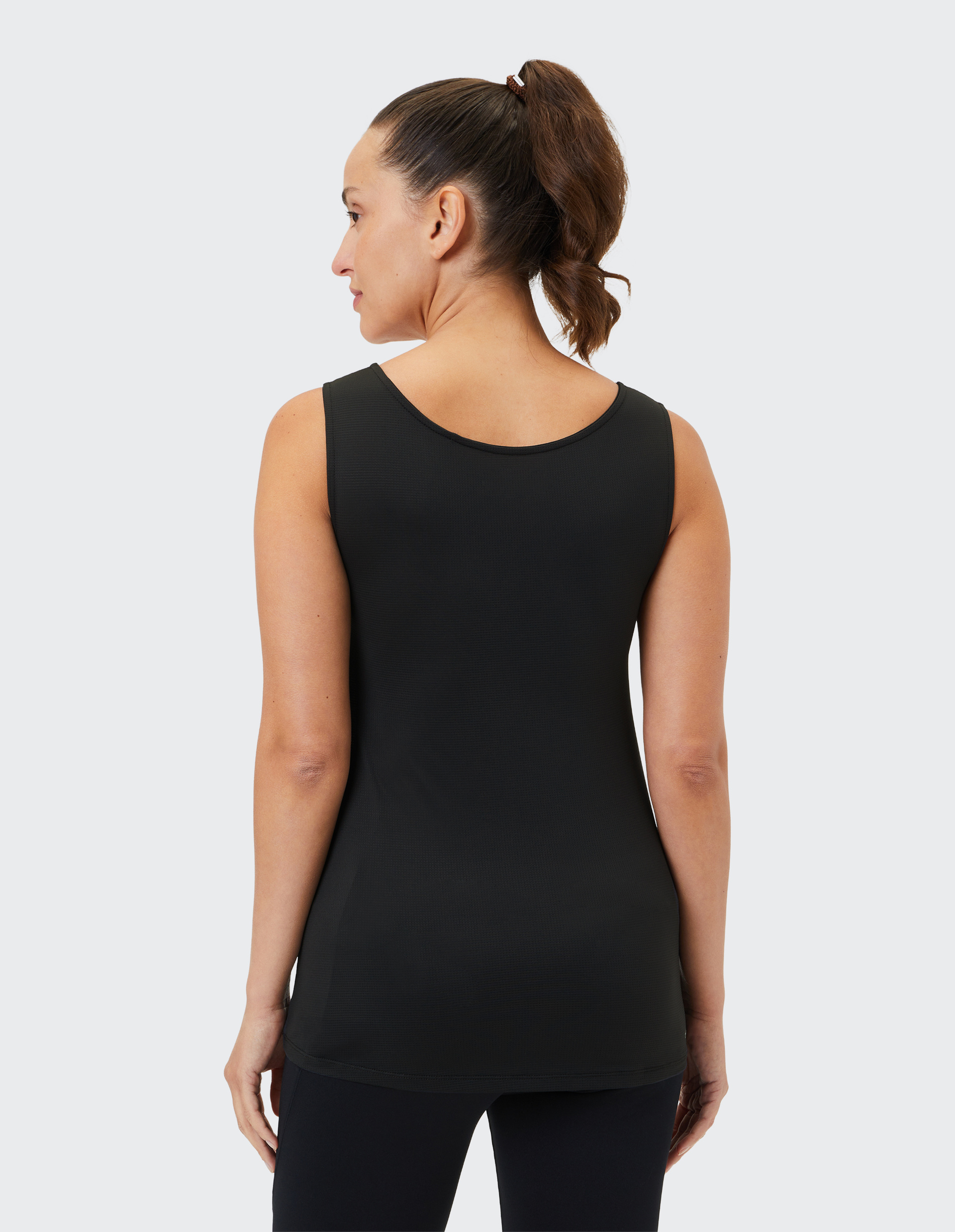 Frau von hinten, trägt schwarzes ärmelloses Top und hat braunes, lockiges Haar im Pferdeschwanz, vor grauem Hintergrund.