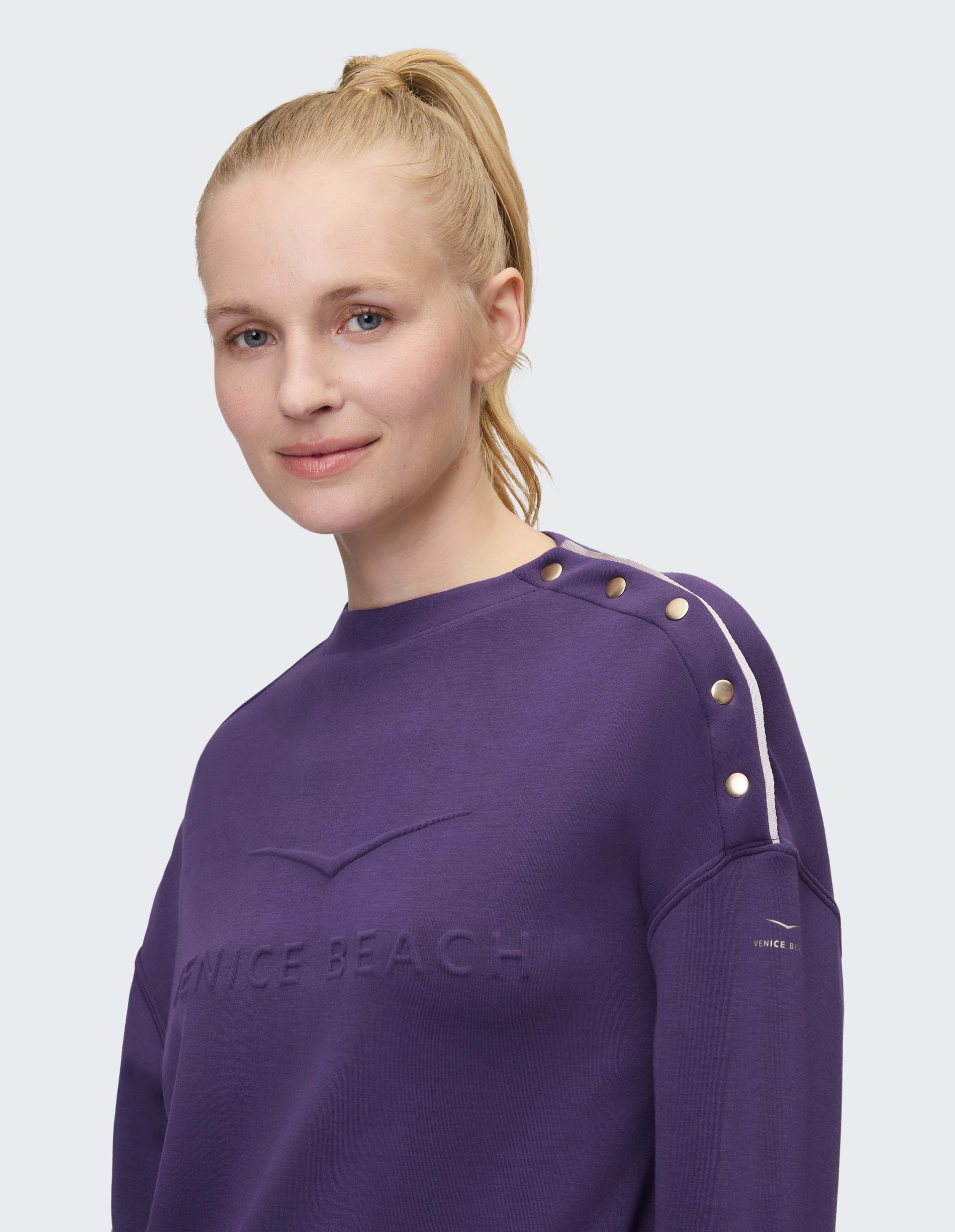 Eine blonde Frau mit Pferdeschwanz trägt einen lila Pullover mit Aufschrift "Venice Beach" und Knöpfen auf der Schulter.