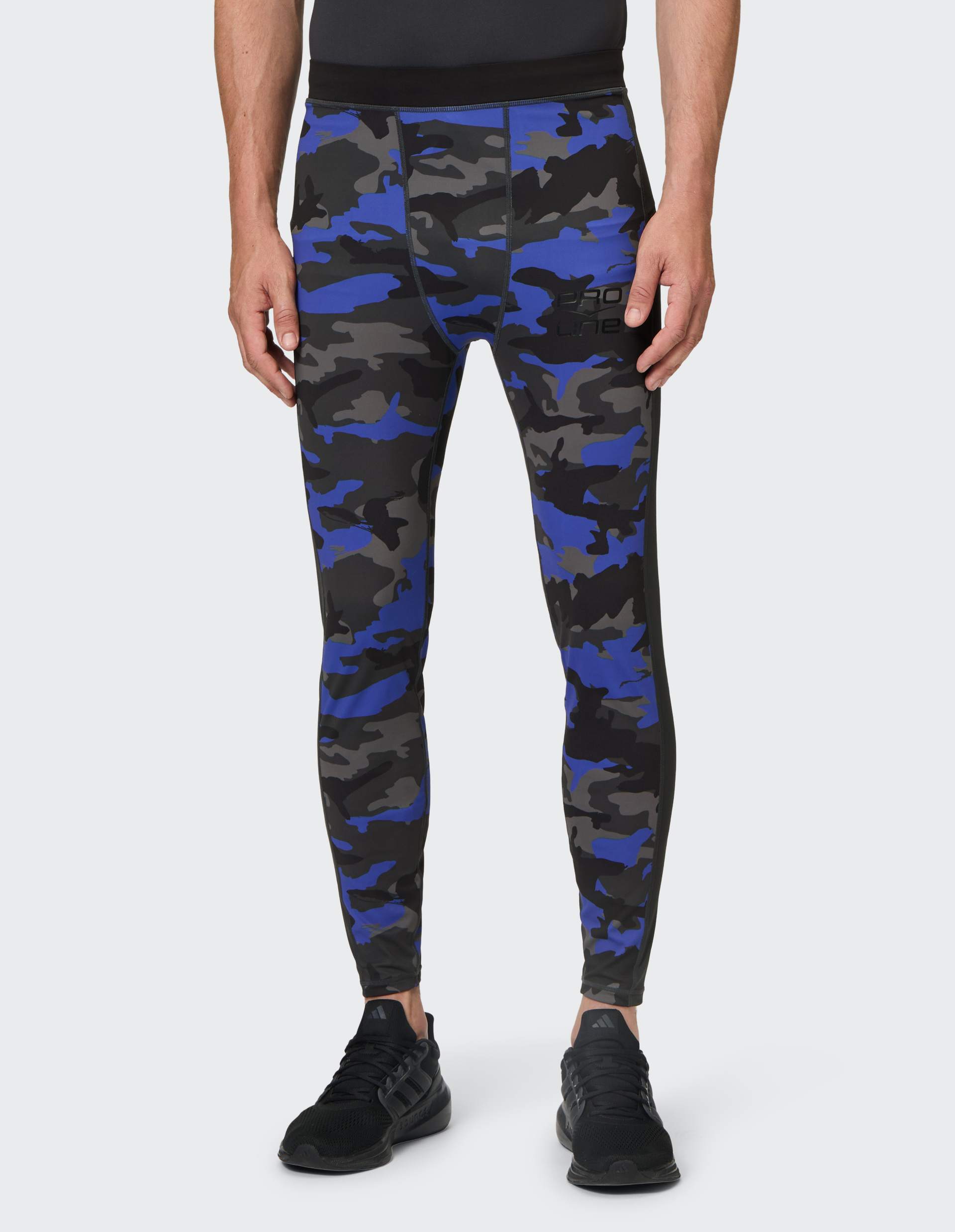 Person trägt eine eng anliegende Leggings mit blau-schwarz-grauem Camouflage-Muster und schwarze Sportschuhe vor grauem Hintergrund.
