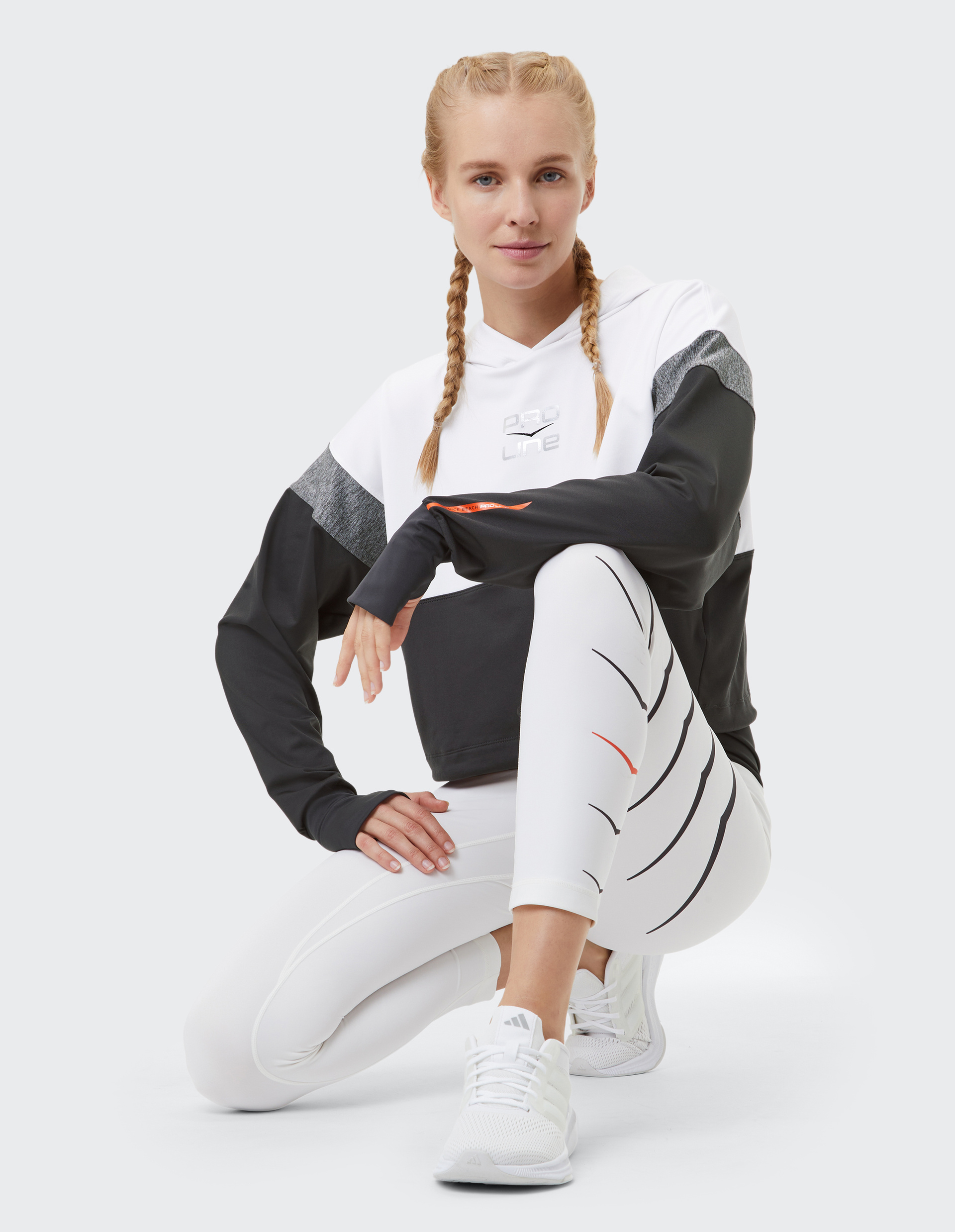 Frau mit geflochtenem Haar kniet in sportlicher Kleidung: schwarz-weißer Hoodie, weiße Leggings mit Streifen und weißen Sneakers.