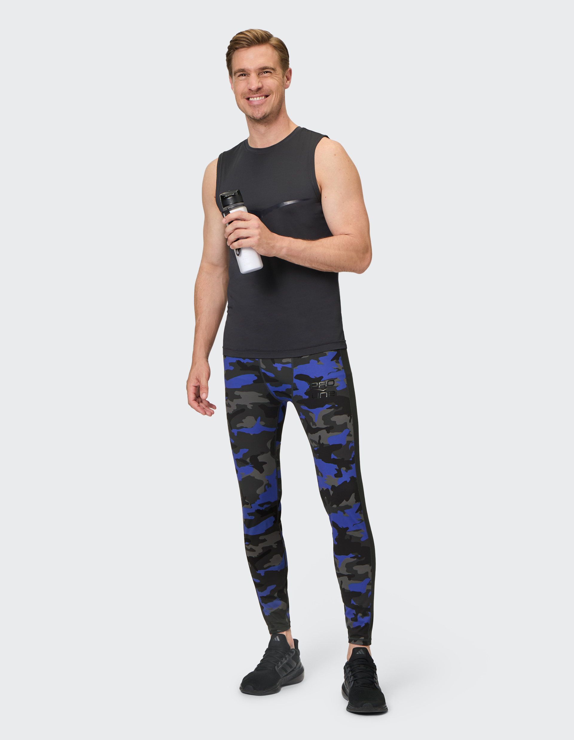 Lächelnder Mann in schwarzem ärmellosen Shirt und blauer Tarn-Leggings hält Trinkflasche vor grauem Hintergrund.