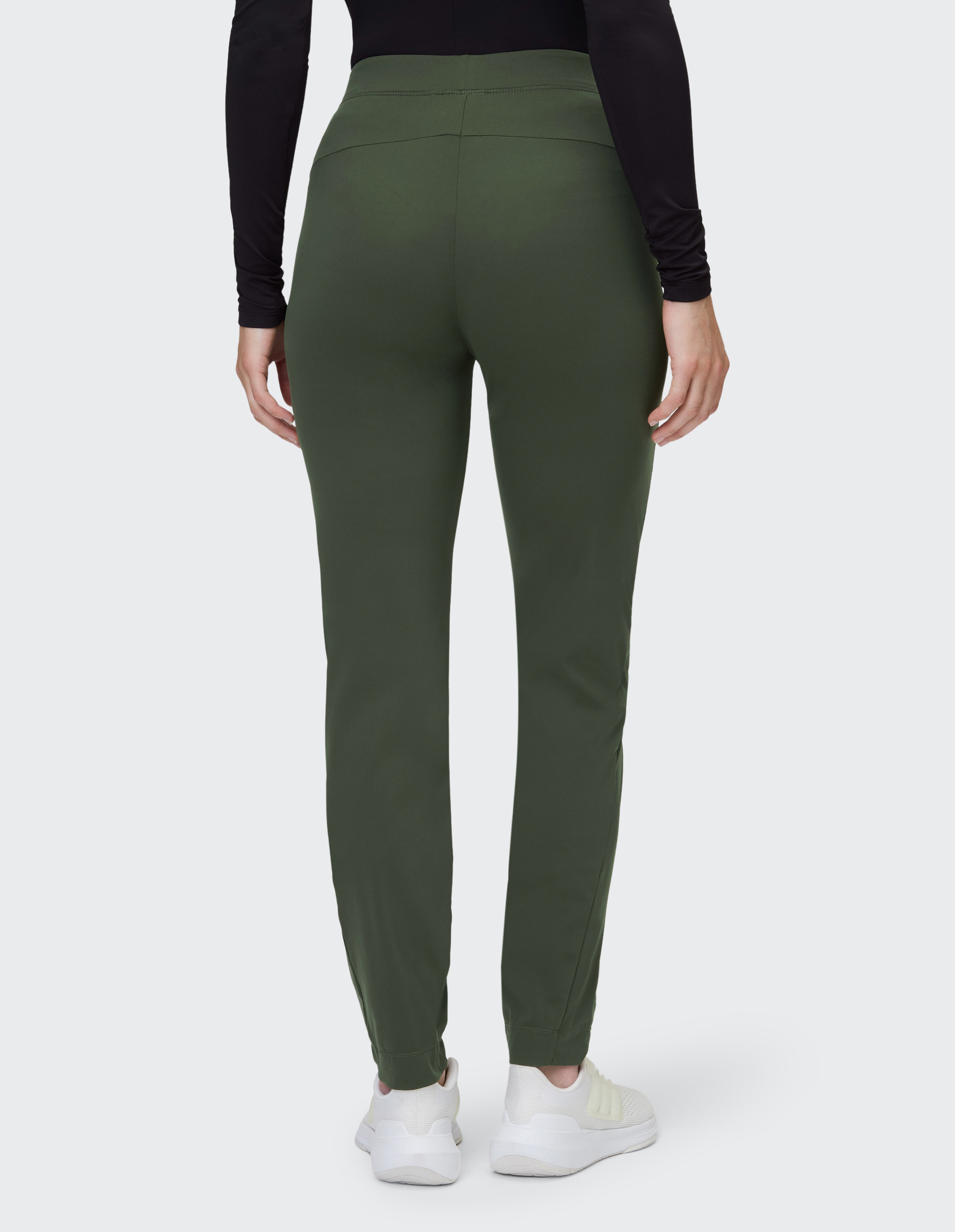 Eine Person trägt grüne, eng anliegende Hosen und weiße Sneakers, fotografiert von hinten, mit schwarzem Oberteil.