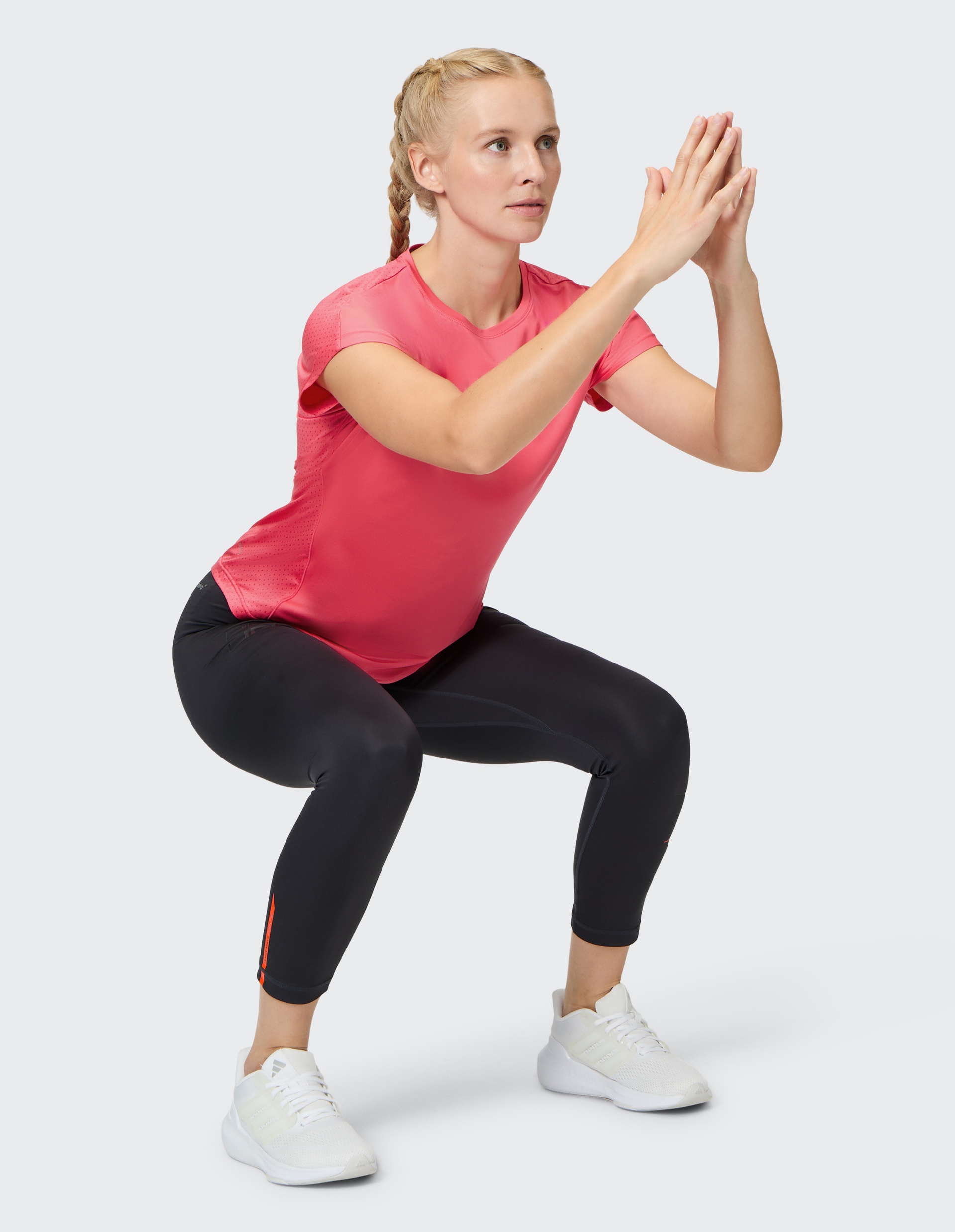 Eine Frau in einem rosa T-Shirt und schwarzen Leggings macht eine Kniebeuge, die Hände sind gefaltet, vor einem neutralen Hintergrund.