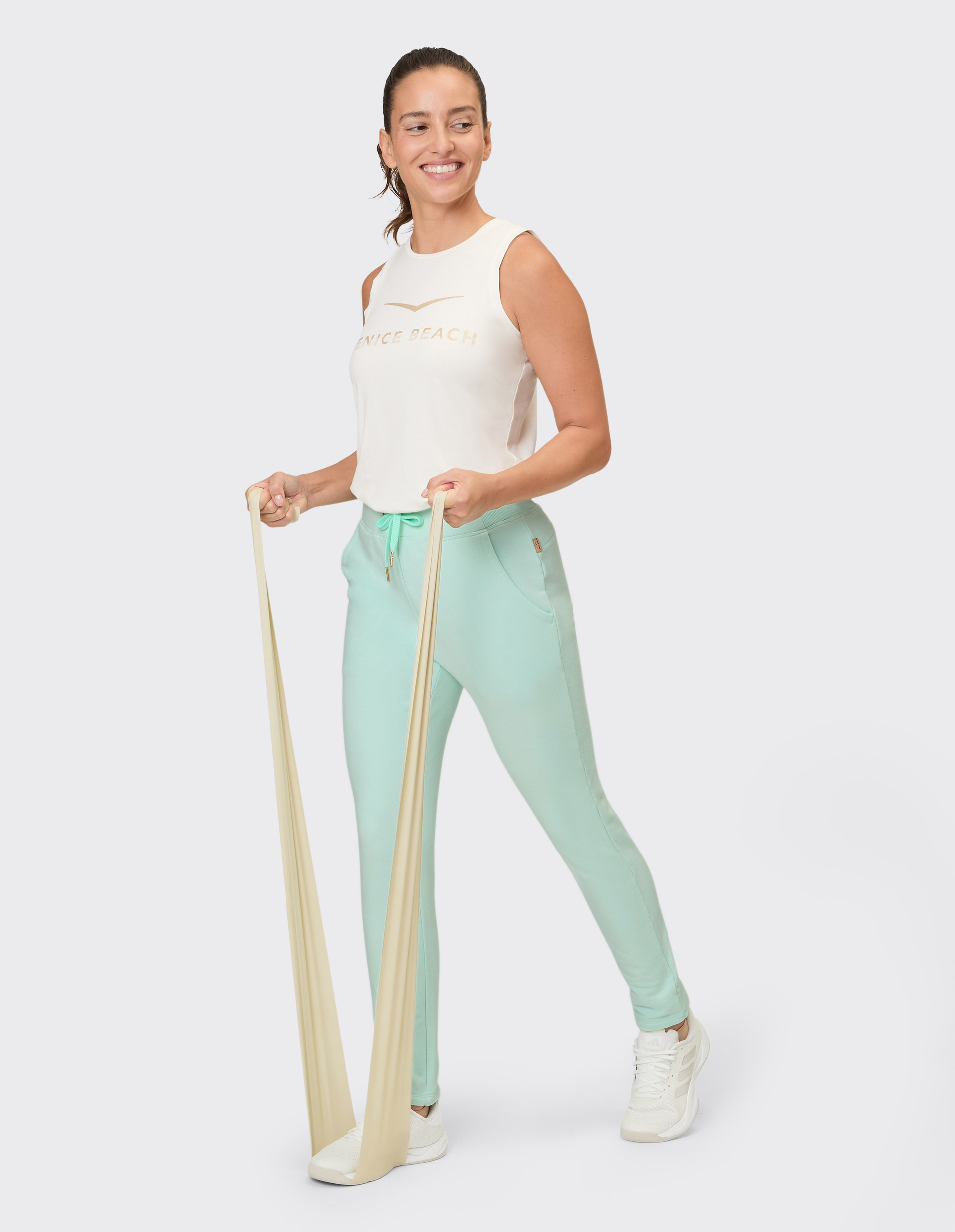 Eine Frau in mintgrüner Trainingshose und einem cremefarbenen Top trainiert mit einem beigefarbenen Widerstandsband.
