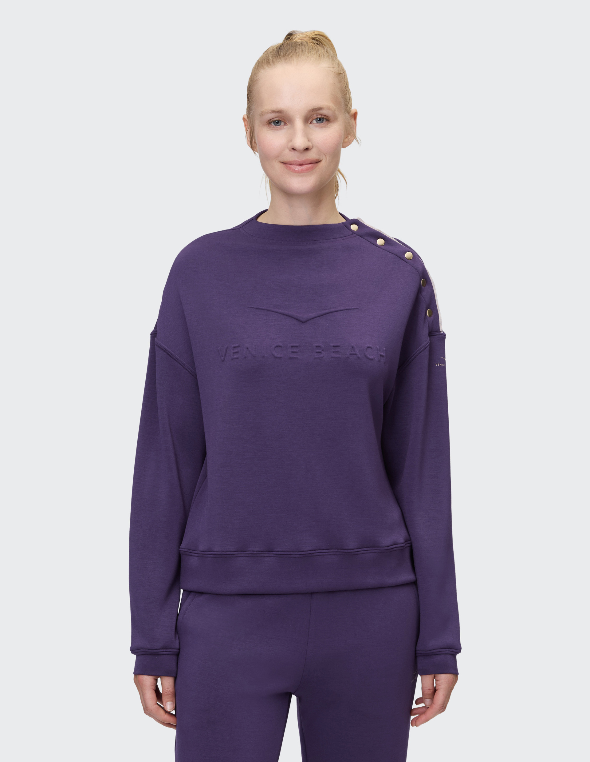 Frau in lila Sweatshirt mit "Venice Beach"-Prägung und Metallic-Knöpfen an der Schulter, lächelt dezent in die Kamera.