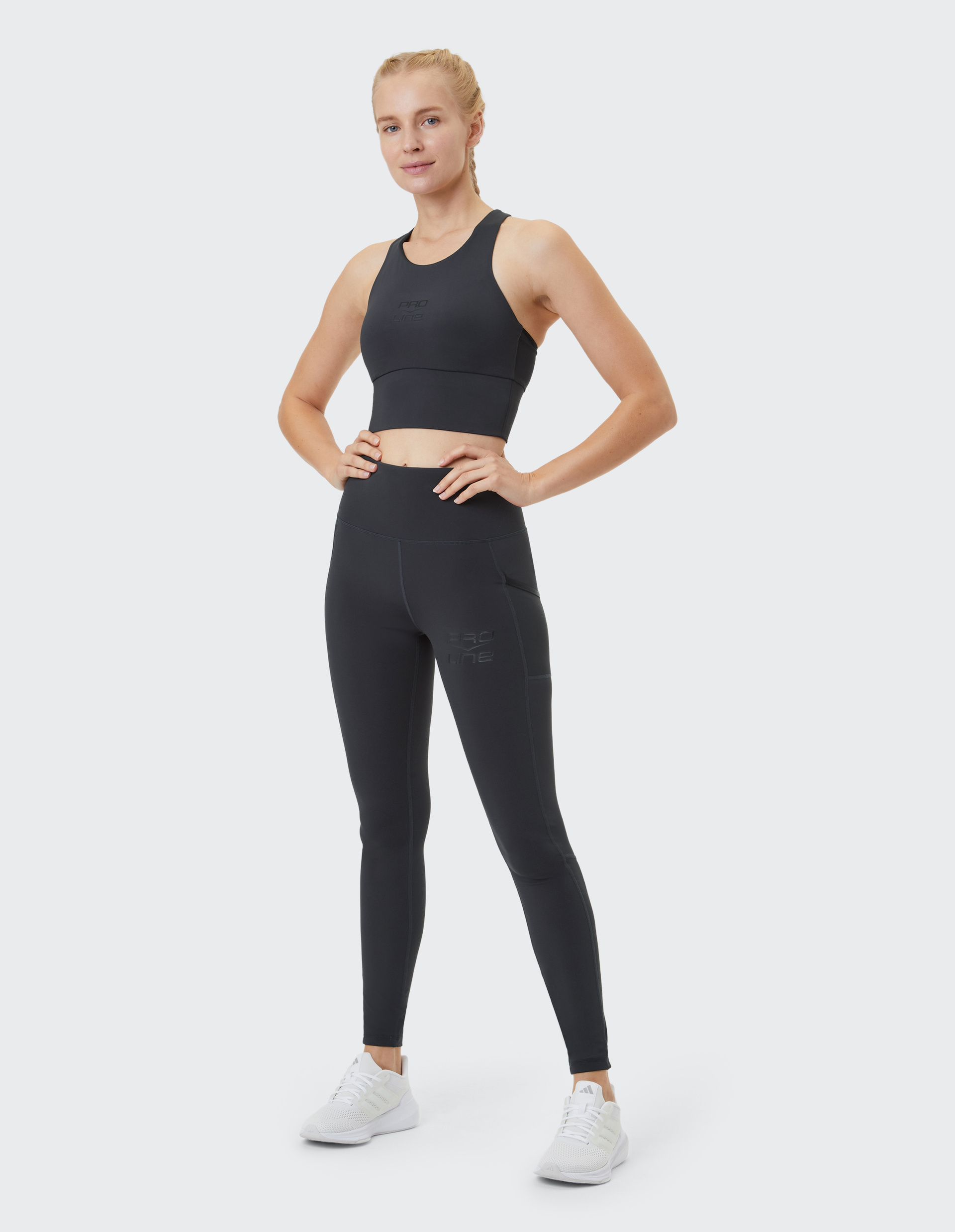 Eine Frau in schwarzem Sport-BH und Leggings steht lächelnd da, die Arme in die Hüften gestützt, auf weißem Hintergrund.