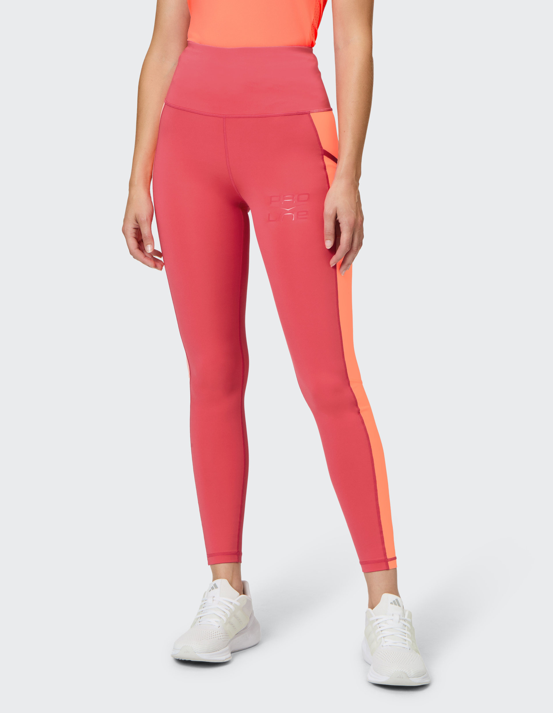 Frau in pinker Sportleggings mit orangefarbenem Streifen und weißem Logo, kombiniert mit weißen Turnschuhen auf hellem Hintergrund.