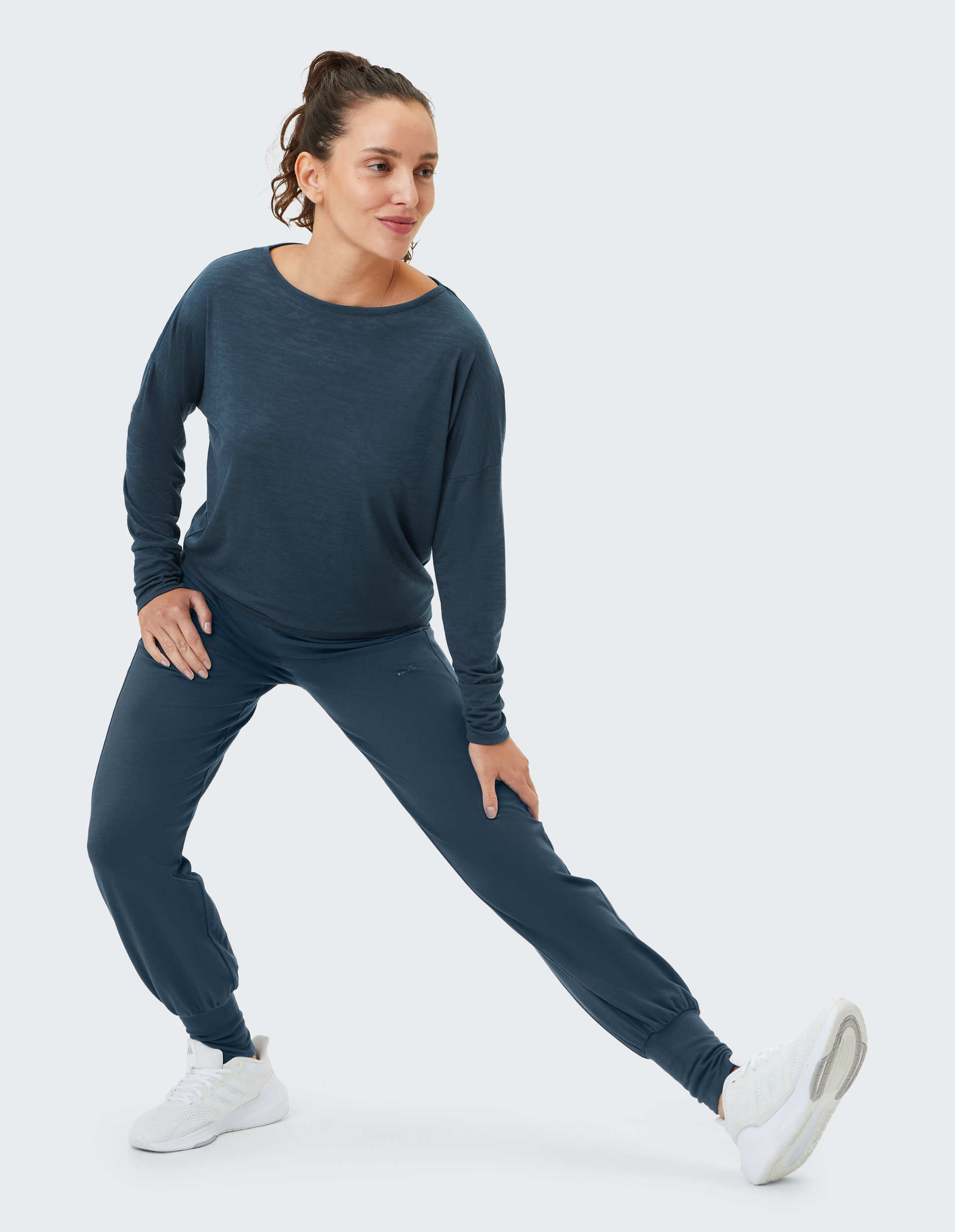 Eine Frau in dunkelblauem Sportoutfit macht im Studio einen Seitenausfallschritt und trägt weiße Turnschuhe.