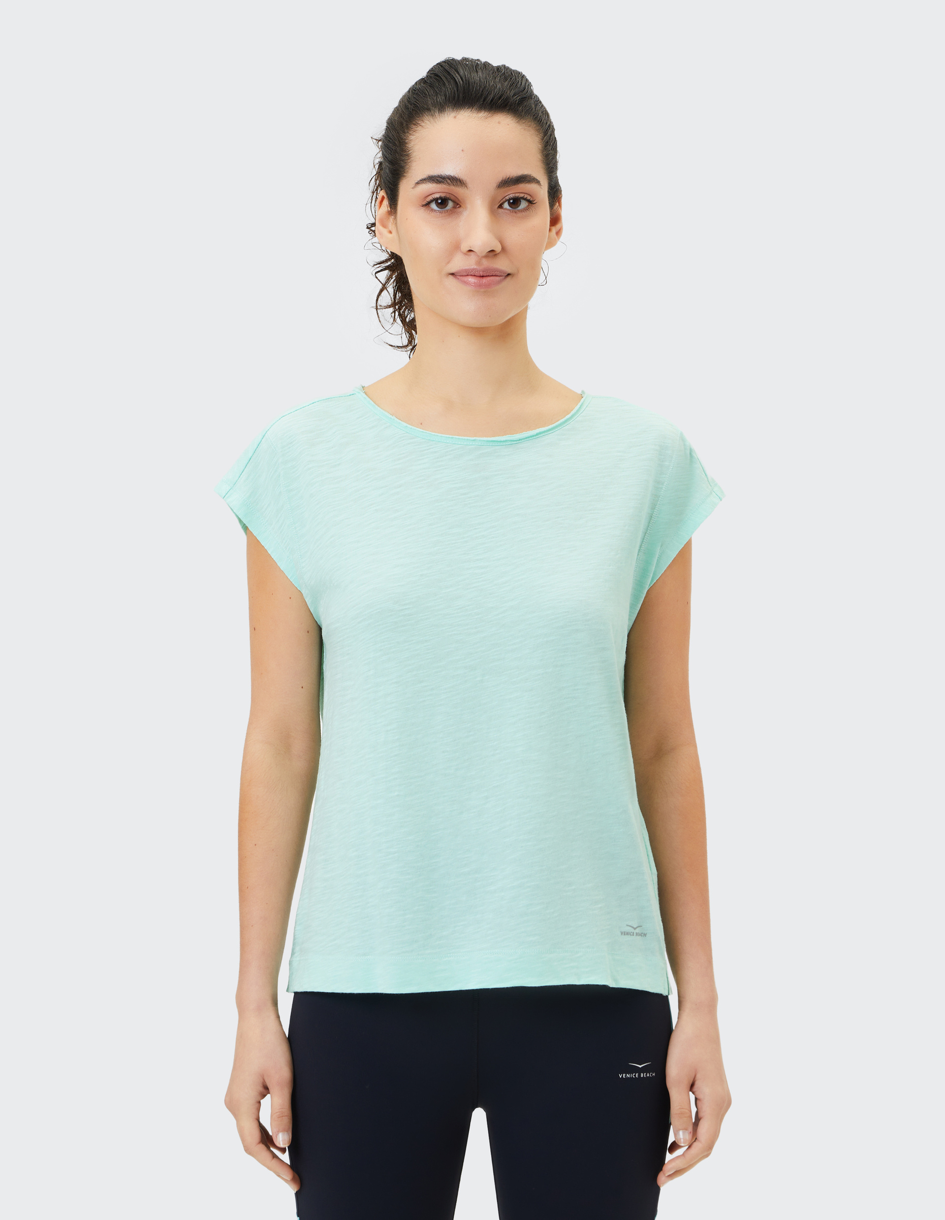 Frau mit lockigem Haar trägt ein mintgrünes T-Shirt und schwarze Leggings, steht lächelnd vor einem hellen Hintergrund.