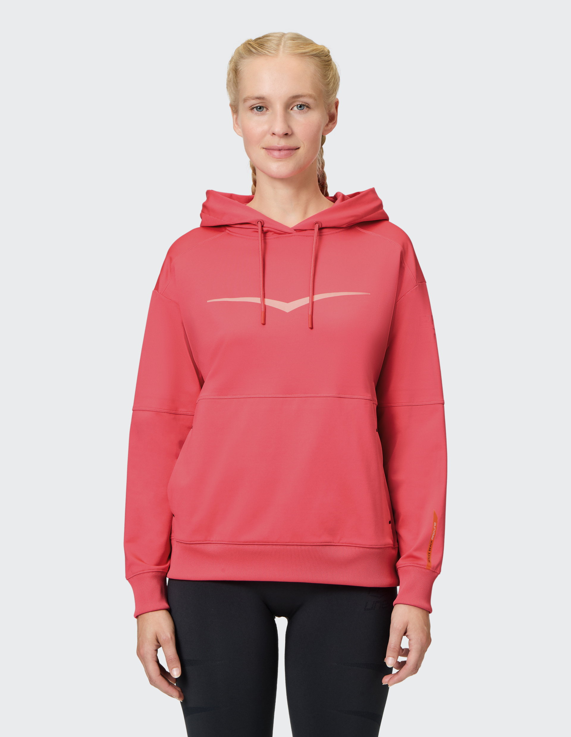 Eine Frau trägt einen roten Kapuzenpullover mit einem dezenten Logo auf der Brust, kombiniert mit schwarzen Leggings vor hellem Hintergrund.