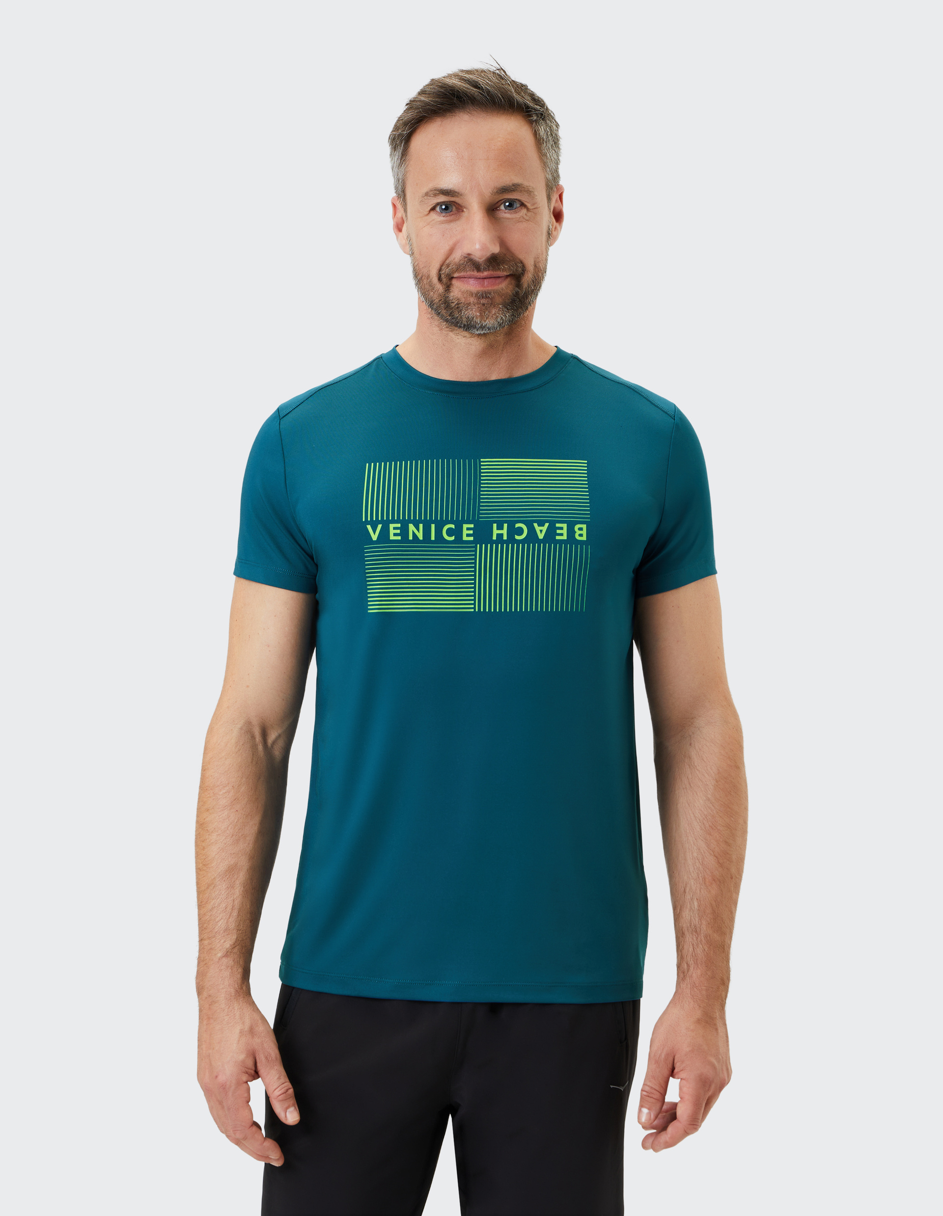 Mann mit Bart trägt ein blaues T-Shirt mit gelbem "Venice Beach"-Schriftzug und schwarzen Hosen, lächelnd vor grauem Hintergrund.