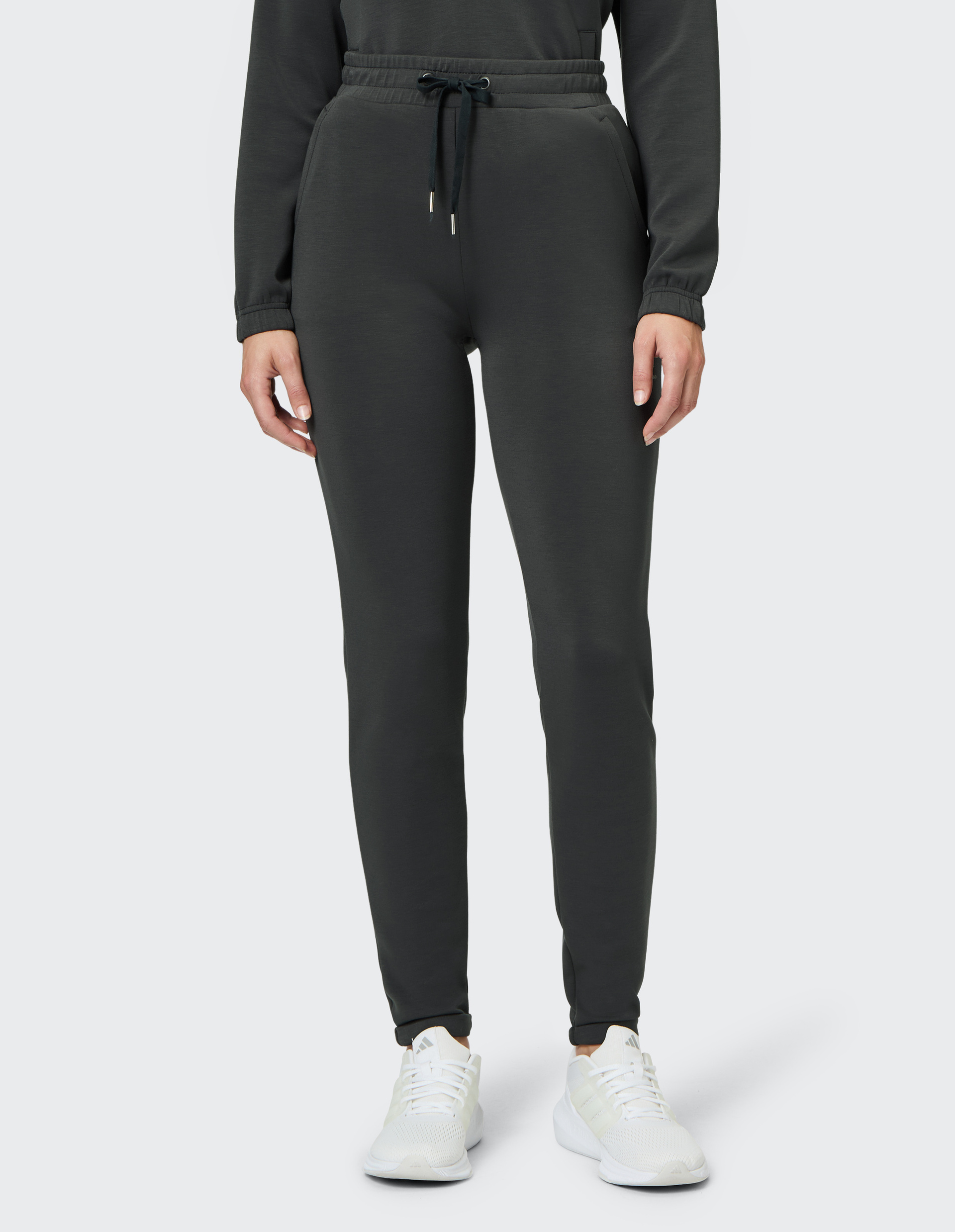 Eine Person trägt eine dunkelgraue Jogginghose mit Kordelzug und weiße Sneakers, stehend vor einem neutralen Hintergrund.
