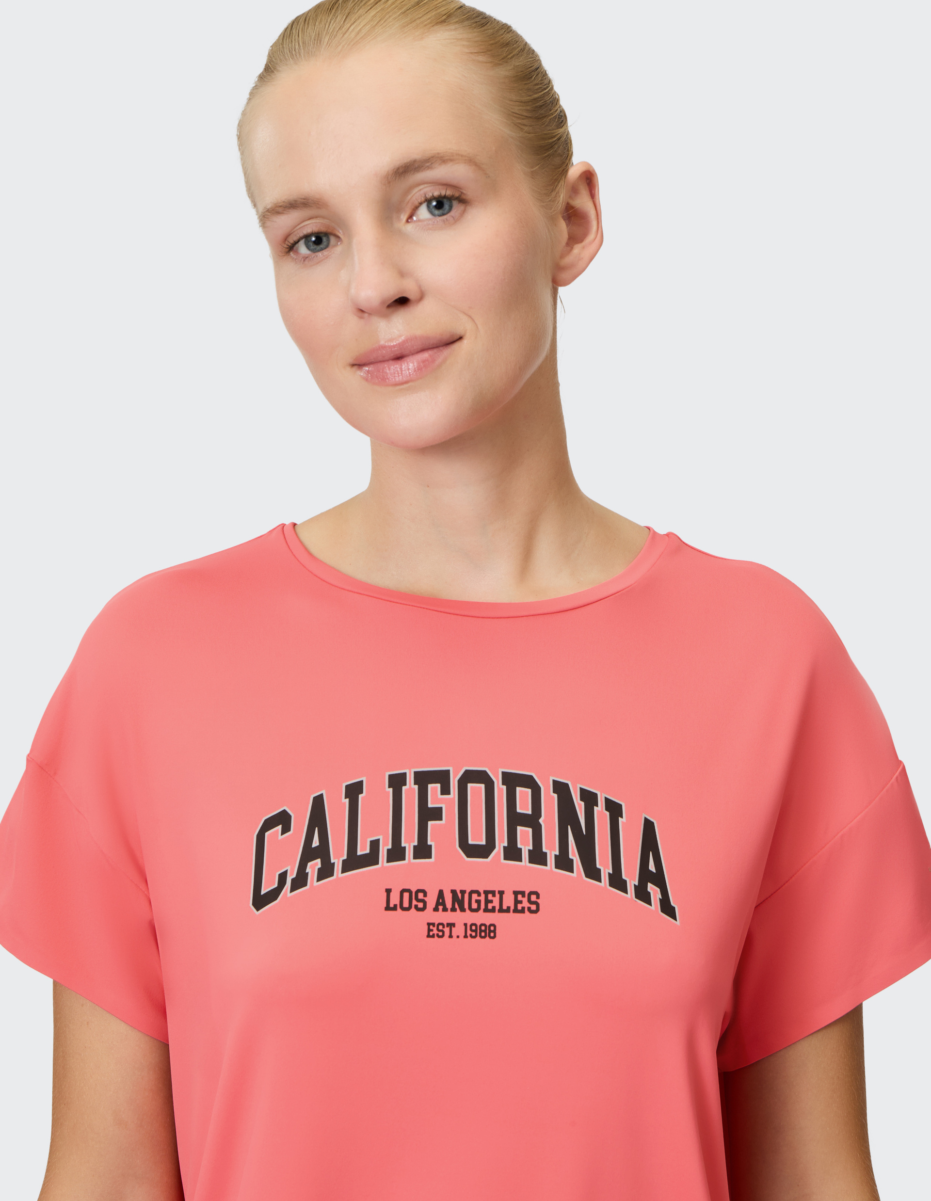 Eine blonde Frau trägt ein pinkes T-Shirt mit der Aufschrift "California Los Angeles Est. 1988" und lächelt leicht.