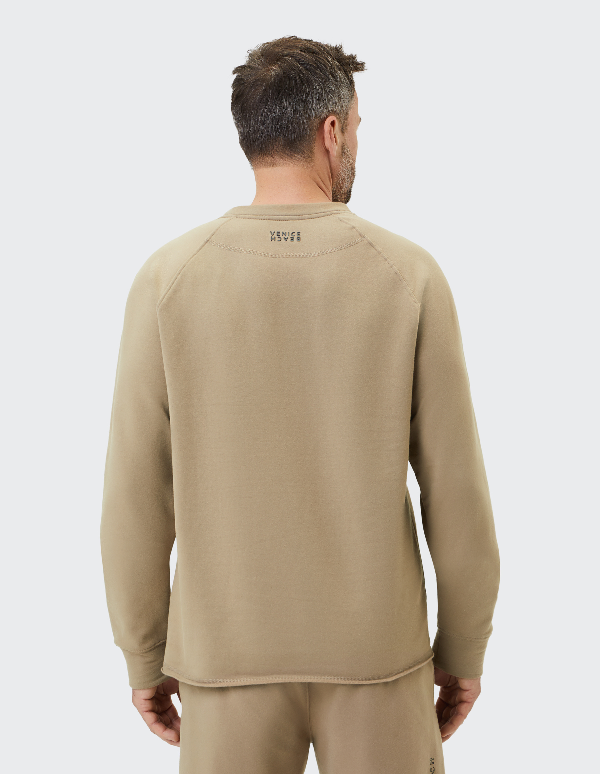 Rückansicht eines Mannes in beige, langärmeligem Sweatshirt und Hose, mit kurzem, dunkelbraunem Haar, auf grauem Hintergrund.