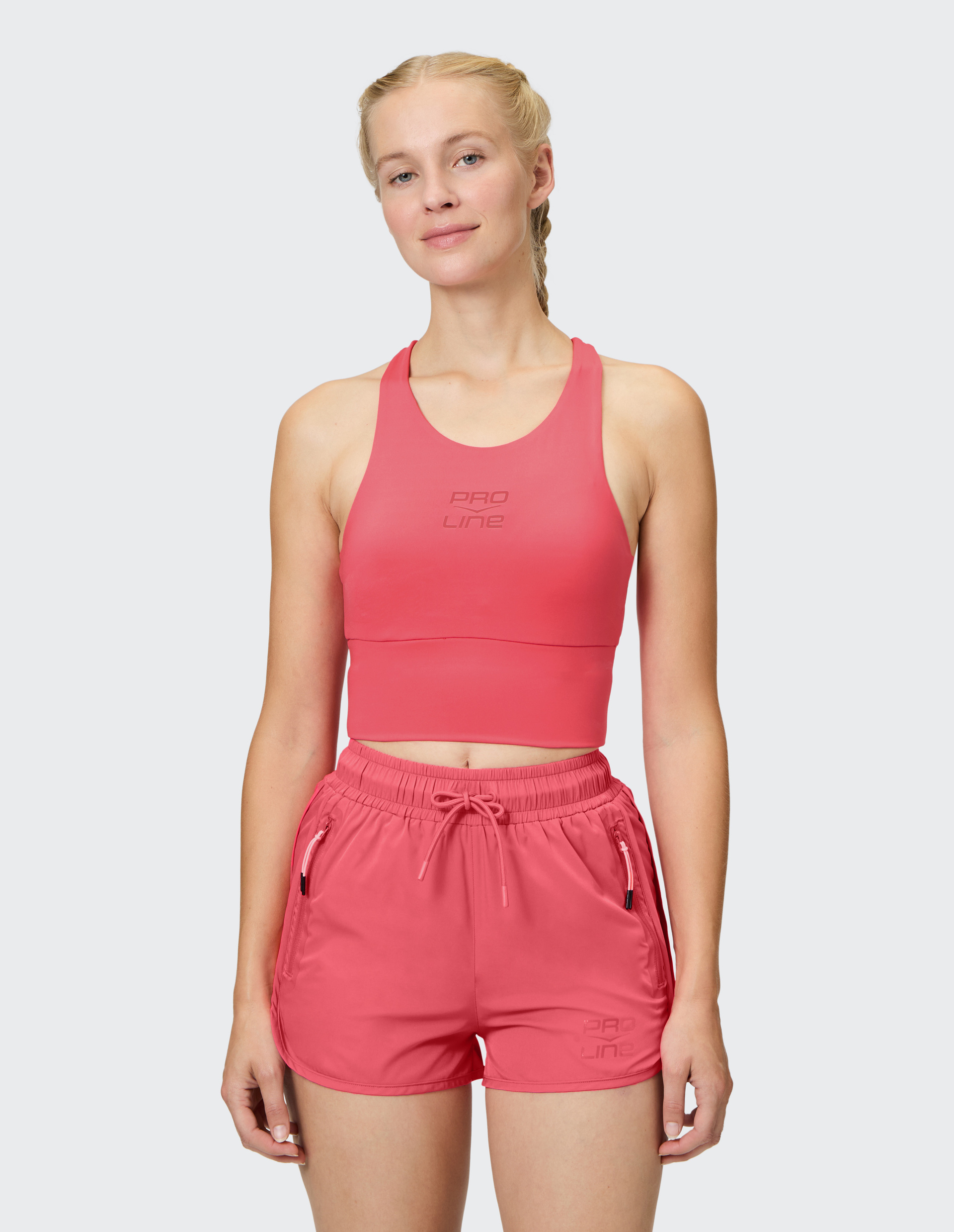 Frau in rosa Sport-BH und kurzen Shorts lächelt leicht, hat blondes Haar in Zöpfen, zeigt Sportkleidung vor neutralem Hintergrund.