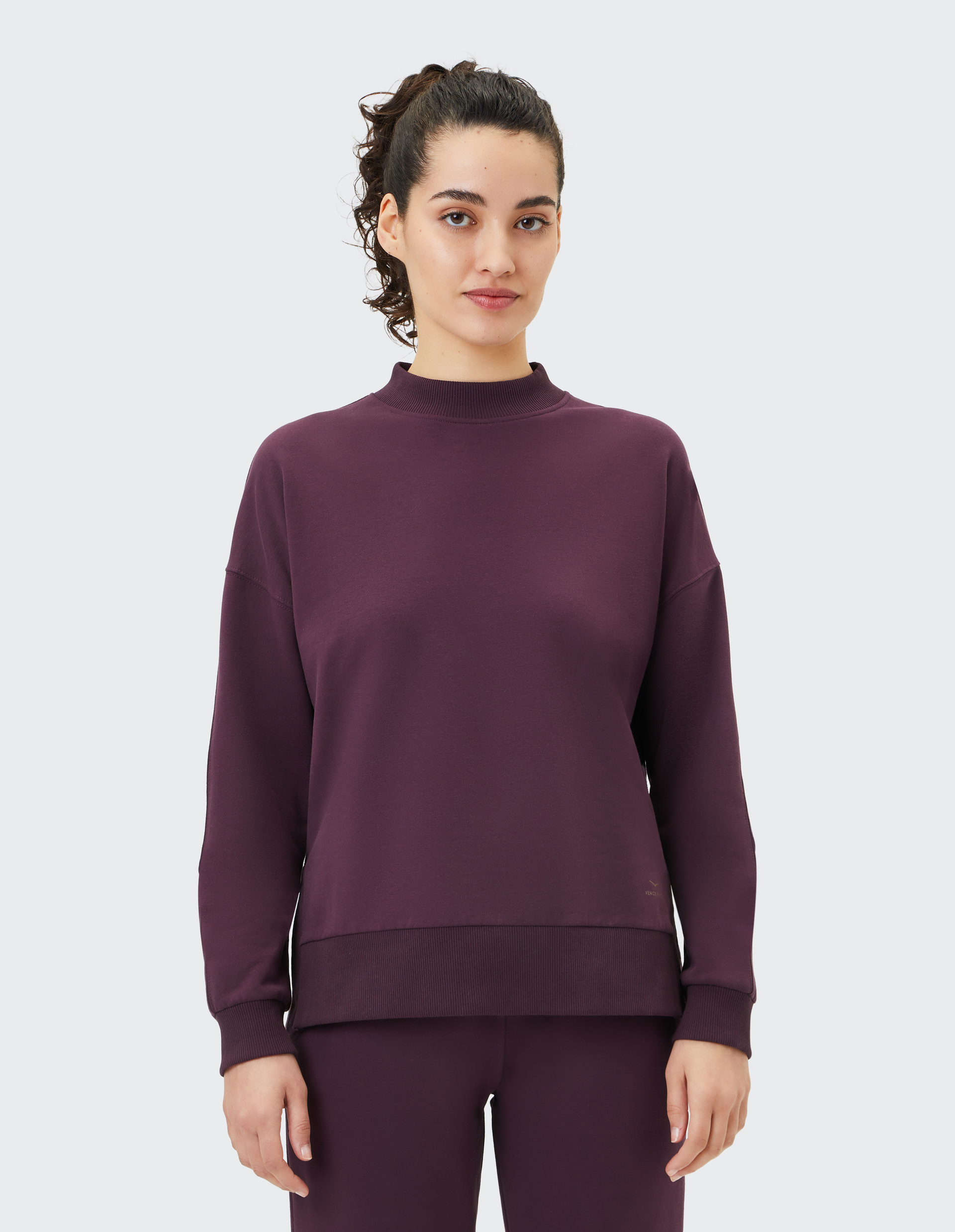 Frau mit lockigem Haar trägt ein bordeauxfarbenes, langärmeliges Sweatshirt und schaut direkt in die Kamera vor neutralem Hintergrund.