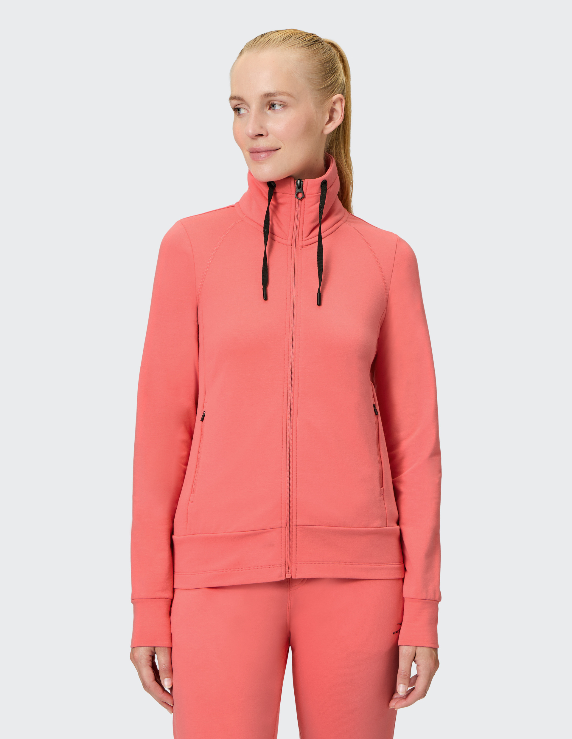 Eine Frau trägt einen rosa Jogginganzug mit hohem Kragen und schwarzen Kordeln, steht seitlich und lächelt leicht.