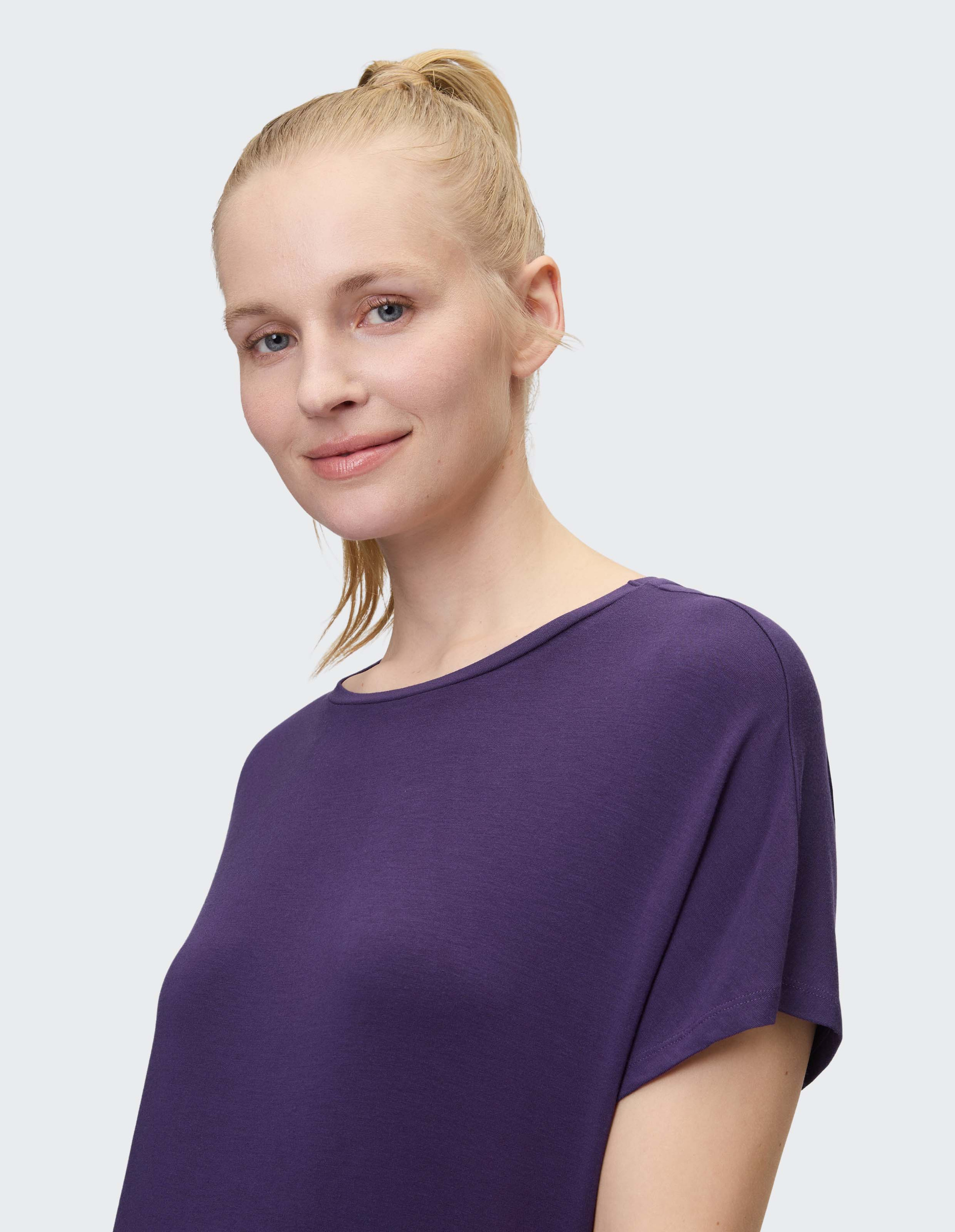 Frau mit blondem Pferdeschwanz und violettem T-Shirt lächelt sanft vor neutralem Hintergrund in die Kamera.