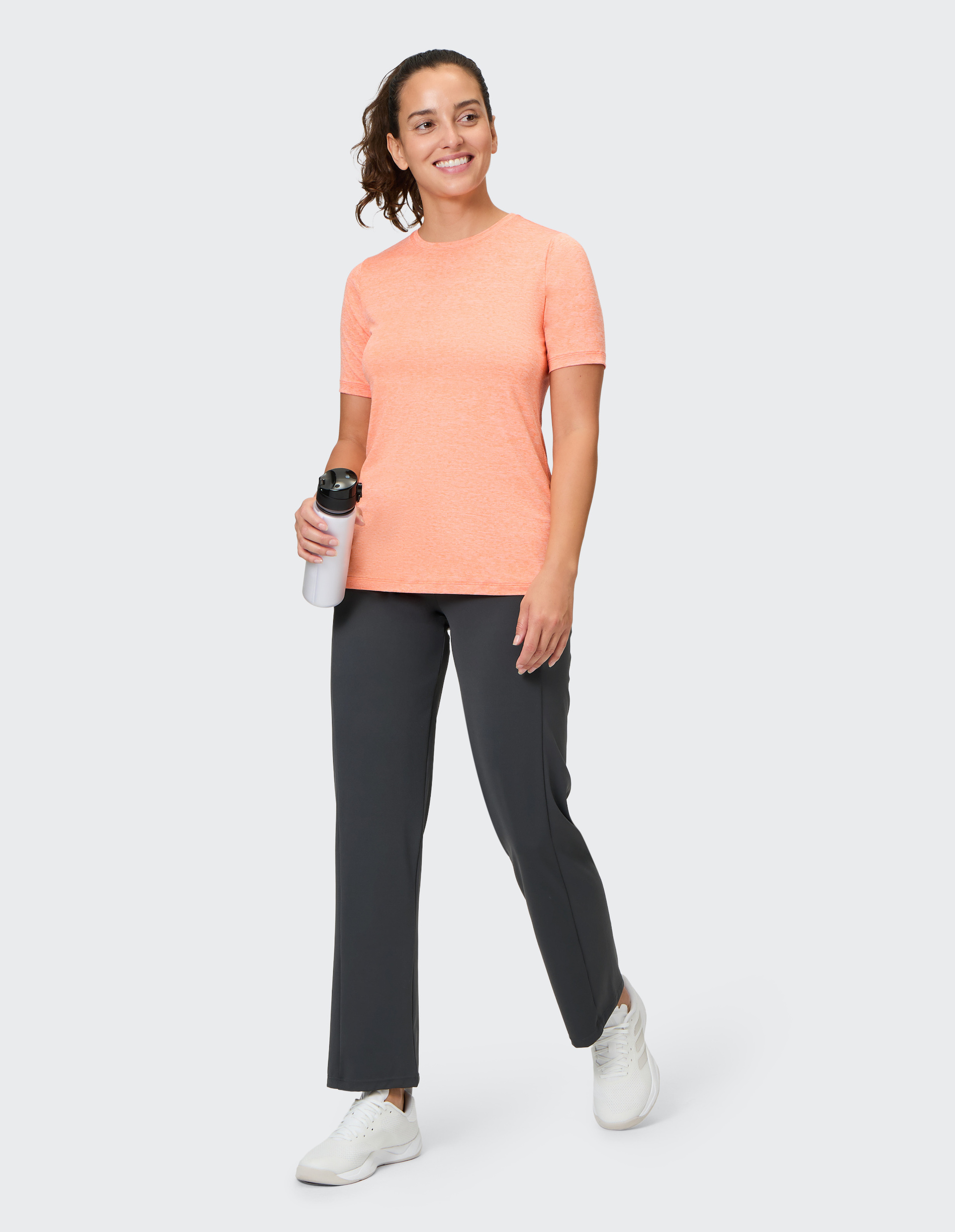 Frau in sportlicher Kleidung mit orangefarbenem T-Shirt hält eine Wasserflasche und geht lächelnd, vor grauem Hintergrund.