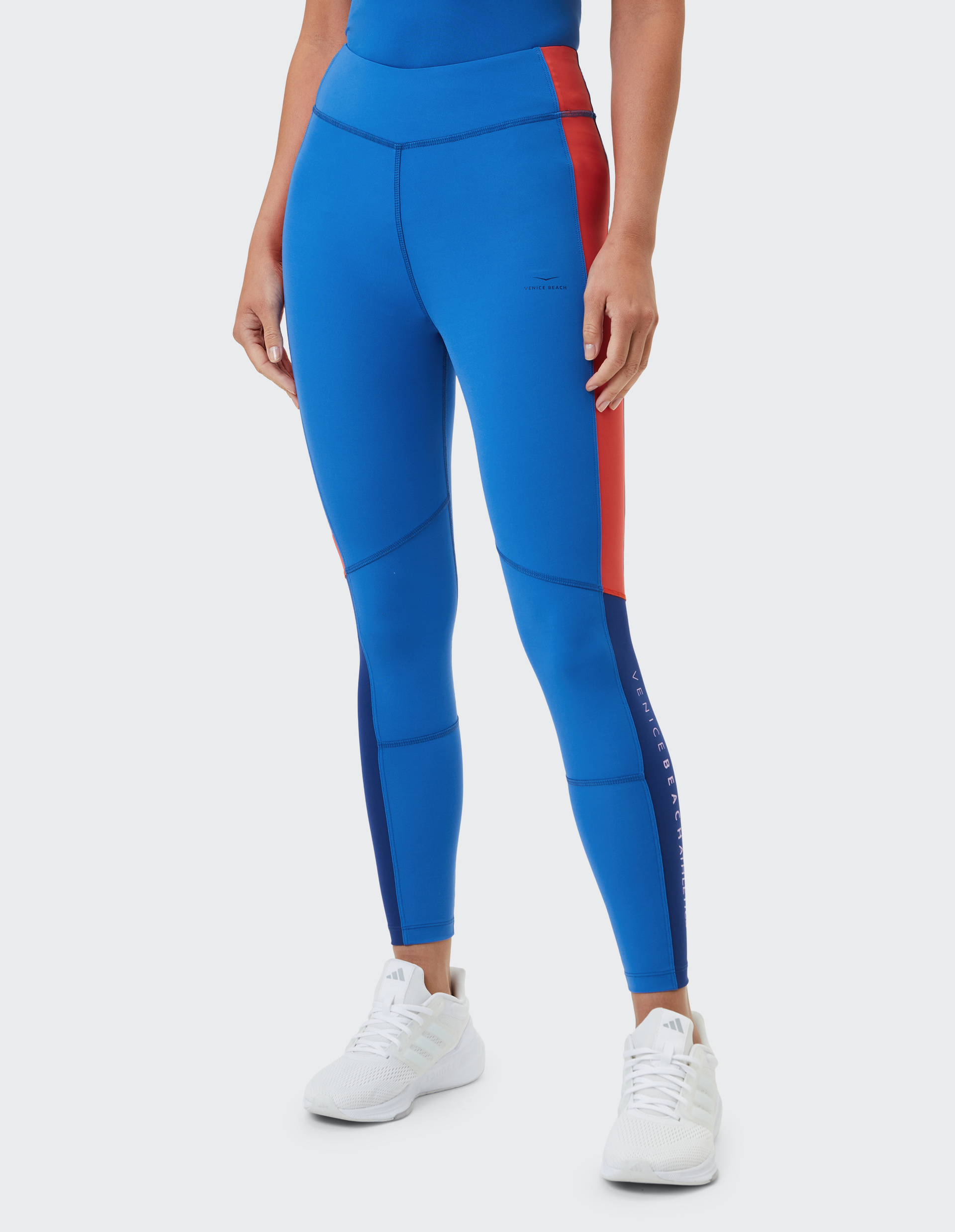 Person trägt blaue Sportleggings mit rotem Streifen, kombiniert mit weißen Turnschuhen auf grau-weißem Hintergrund.