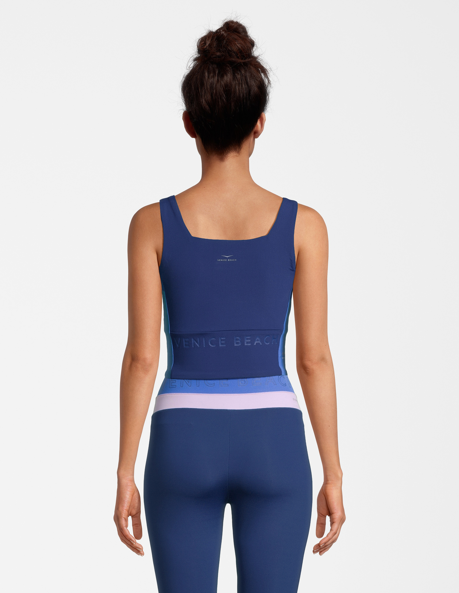 Frau von hinten in blauem Sporttop und Leggings mit VENICE BEACH-Schriftzug auf Rückseite, Haar in Dutt, vor weißem Hintergrund.