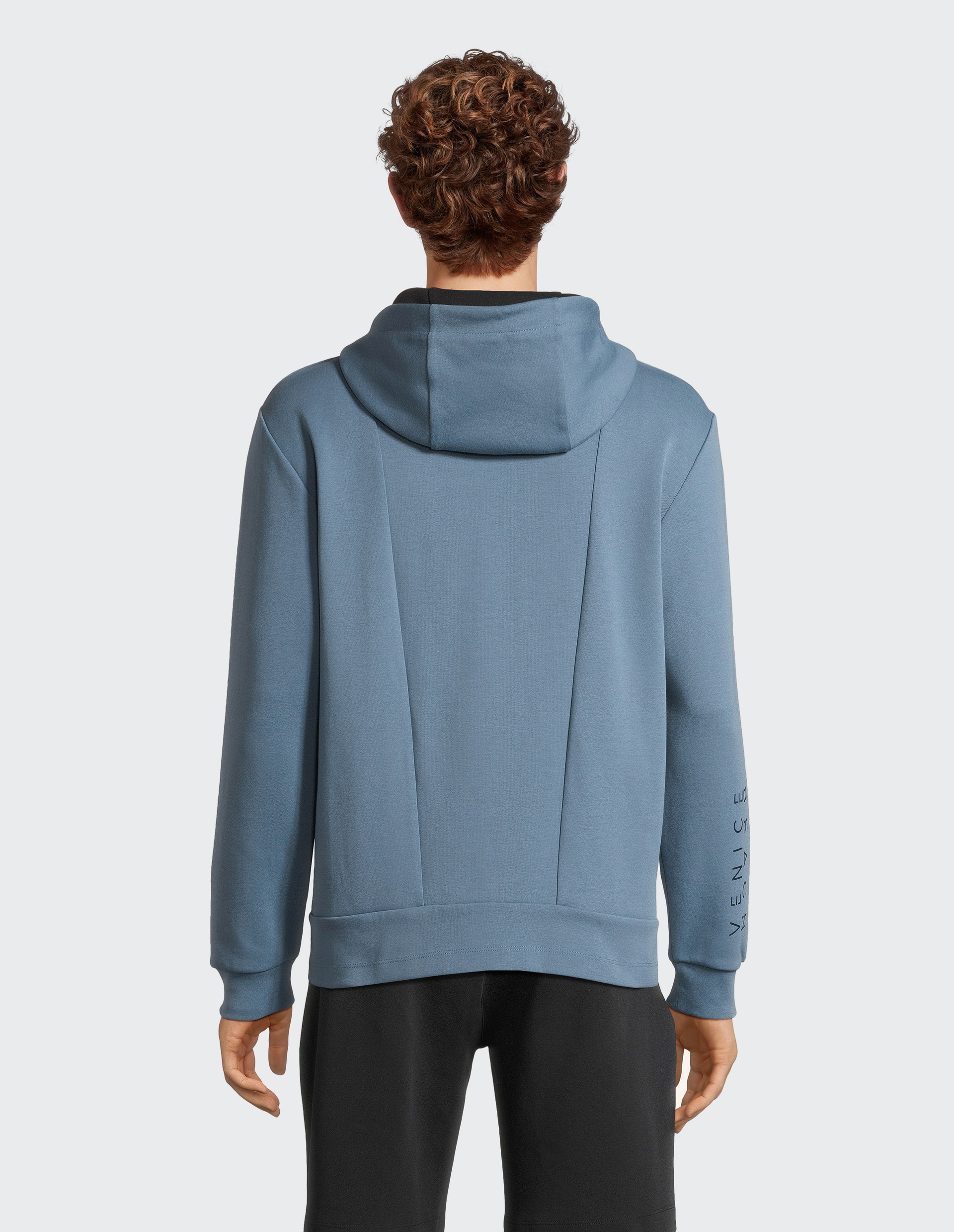 Eine Person mit lockigem Haar trägt einen blauen Hoodie und schwarze Hosen, von hinten gezeigt, auf grauem Hintergrund.