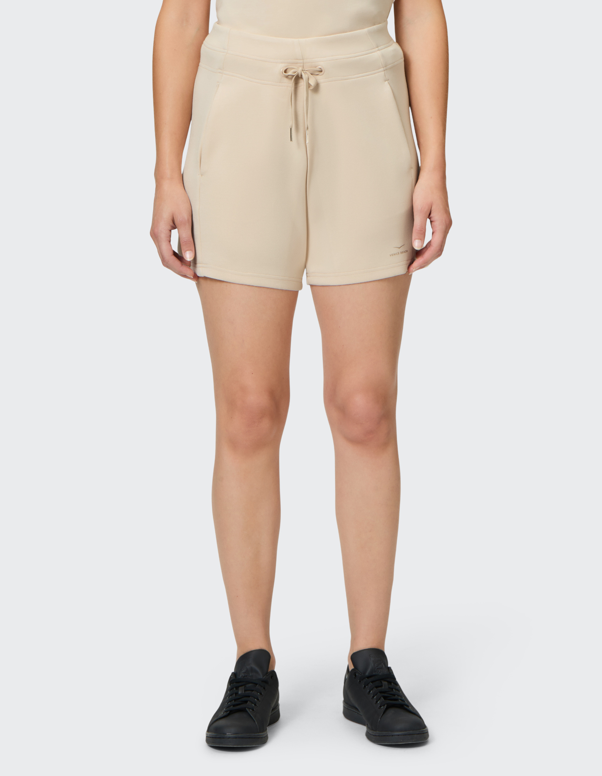 Eine Person trägt beigefarbene Shorts mit Kordelzug und schwarzen Sneakers, stehend auf grauem Hintergrund.