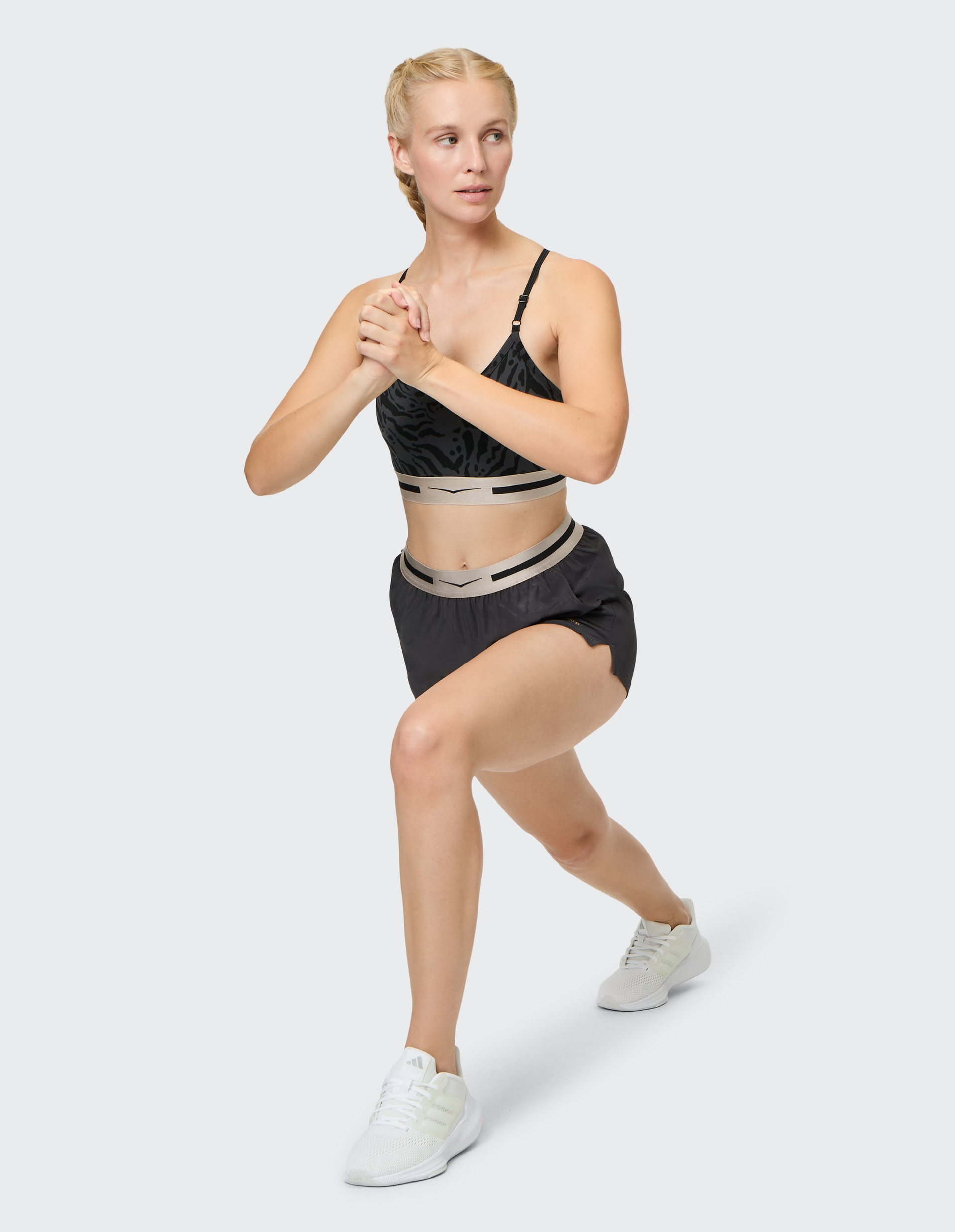 Frau in Sportkleidung macht Ausfallschritt-Übung auf grauem Hintergrund, Hände zusammengenommen, Blick zur Seite.