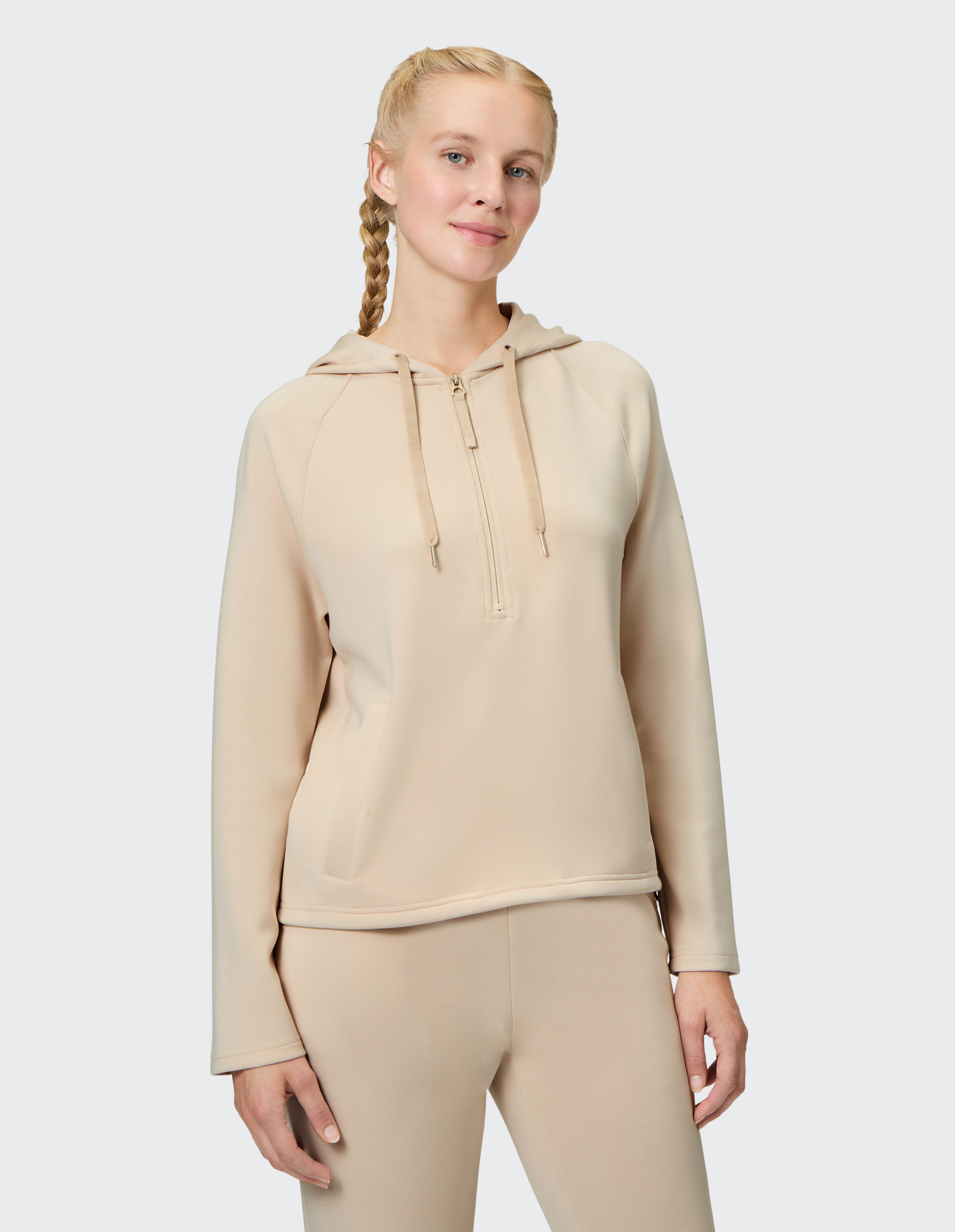 Frau mit einem beigen Kapuzenpullover und Zopffrisur steht vor neutralem Hintergrund und schaut freundlich in die Kamera.