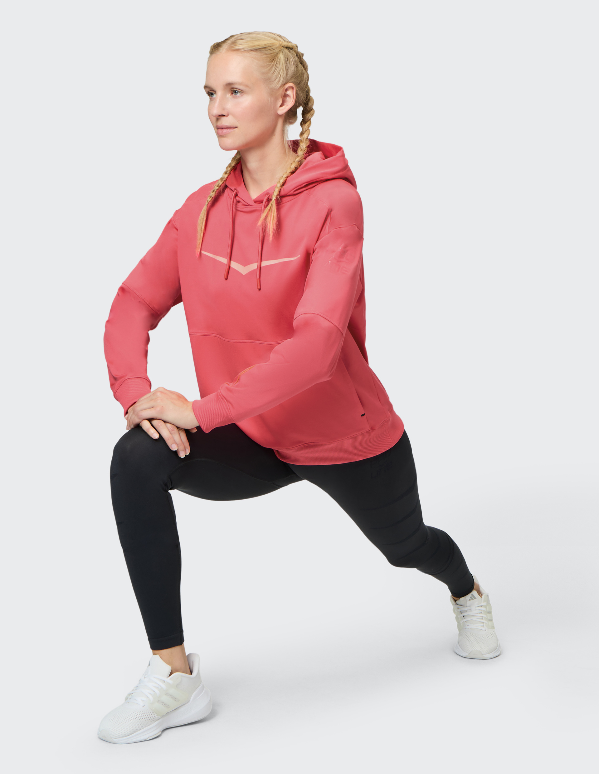 Frau in rosa Hoodie und schwarzen Leggings macht Dehnübung mit Ausfallschritt, Zöpfe und weiße Sneakers auf hellem Hintergrund.