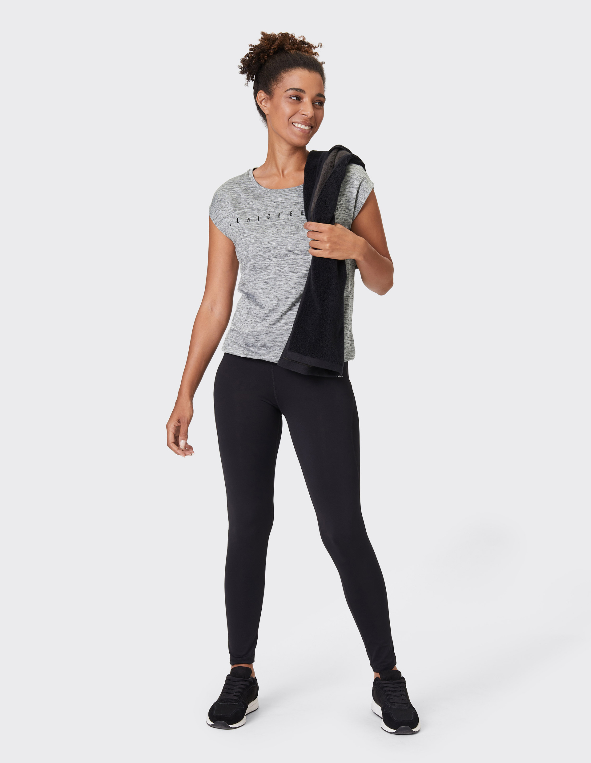 Sportlich gekleidete Frau mit grauem Shirt und schwarzer Leggings hält Handtuch über der Schulter, lächelt und steht aufrecht.