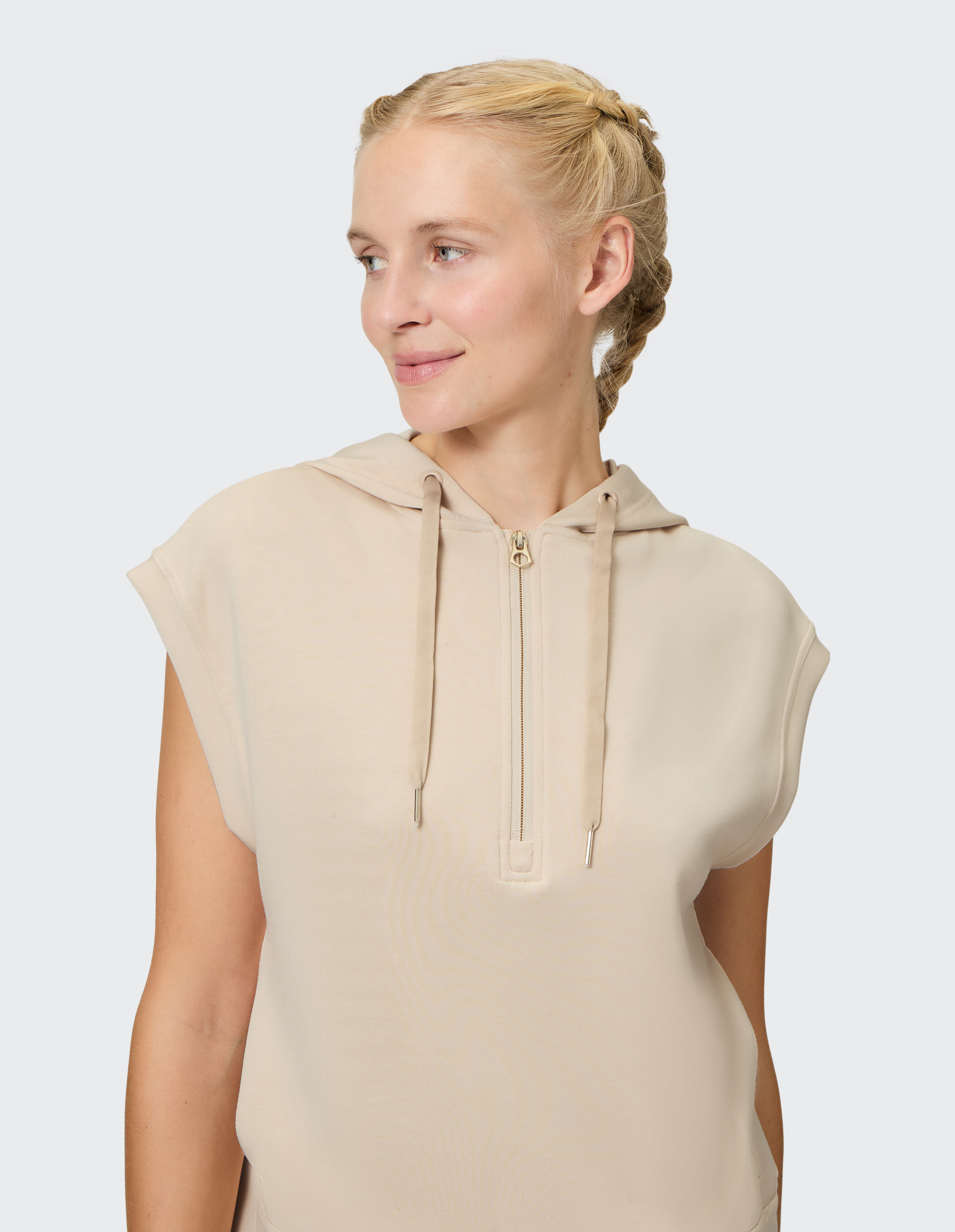 Eine Frau trägt ein ärmelloses, beigefarbenes Hoodie-Kleid mit Zopf, lächelt leicht und blickt seitlich auf grauem Hintergrund.