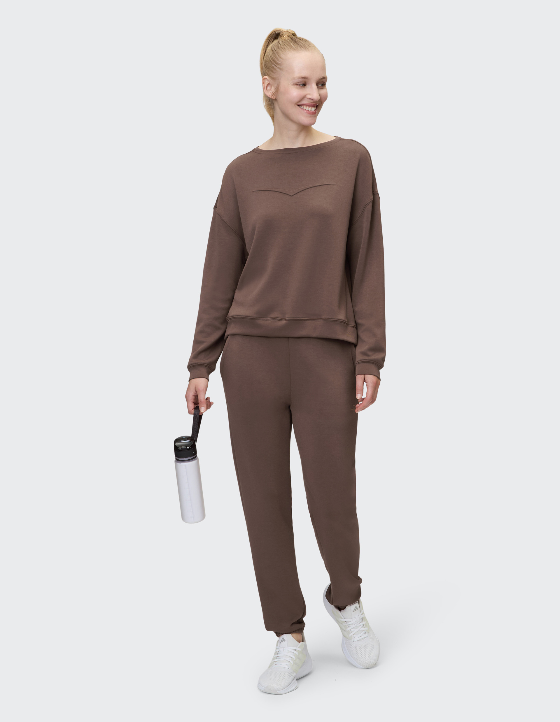 Frau in braunem Freizeitanzug mit entspanntem Schnitt und weißen Turnschuhen, Haare zu einem Zopf gebunden, steht vor grauem Hintergrund.