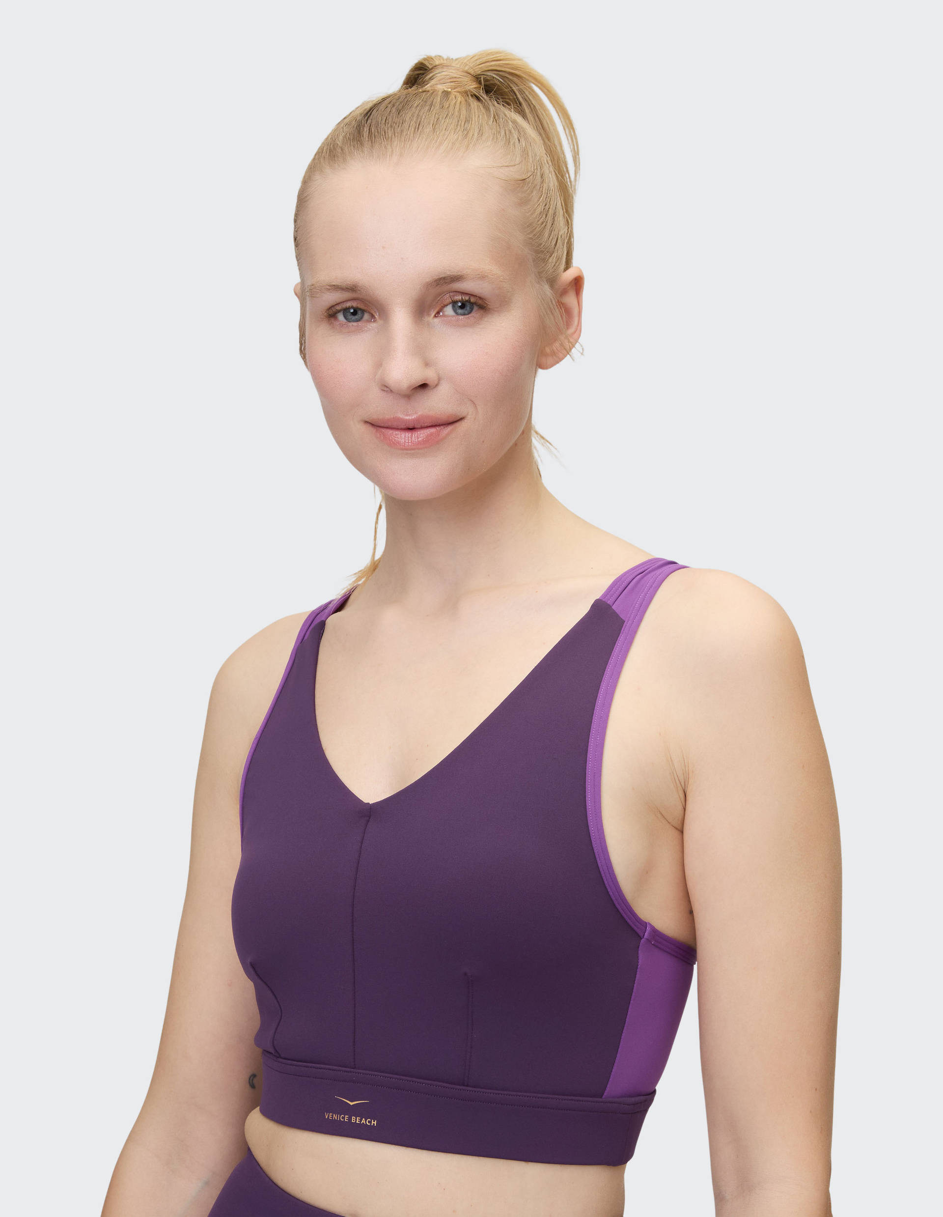 Eine lächelnde Person in einem lila Sporttop mit Zopf steht vor einem neutralen Hintergrund.