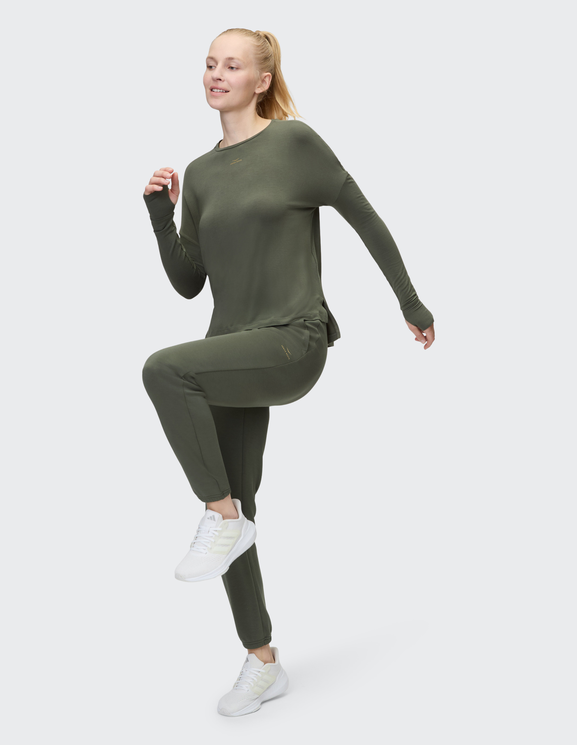 Eine Person trägt ein olivgrünes Langarmshirt und passende Jogginghose vor einem neutralen Hintergrund mit weißen Turnschuhen.